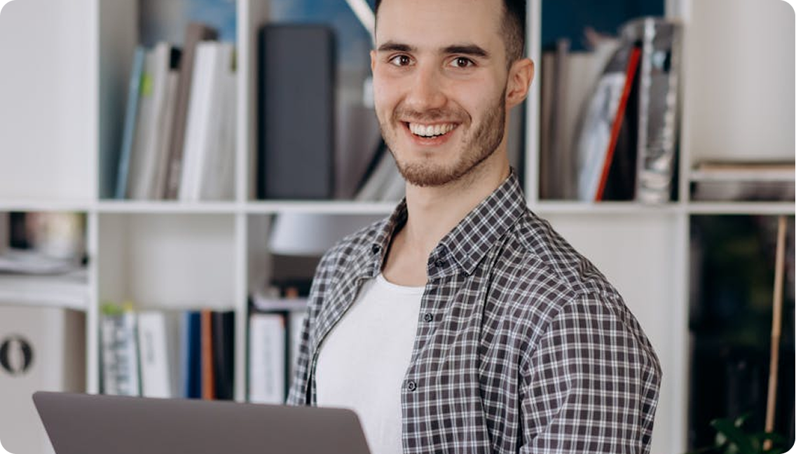 A young man with short hair, a beard, wearing a checkered shirt over a white t-shirt, smiling, standing in an office or study space with shelves filled with books and files behind him.