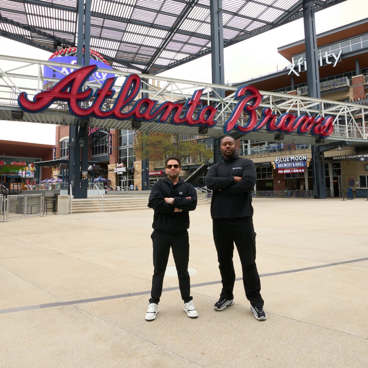 Two men standing in front of the Atlanta Braves stadium sign at Truist Park. The man on the left is wearing sunglasses and a black jacket, while the man on the right is taller with a beard and wearing a black hoodie. Both have their arms crossed.