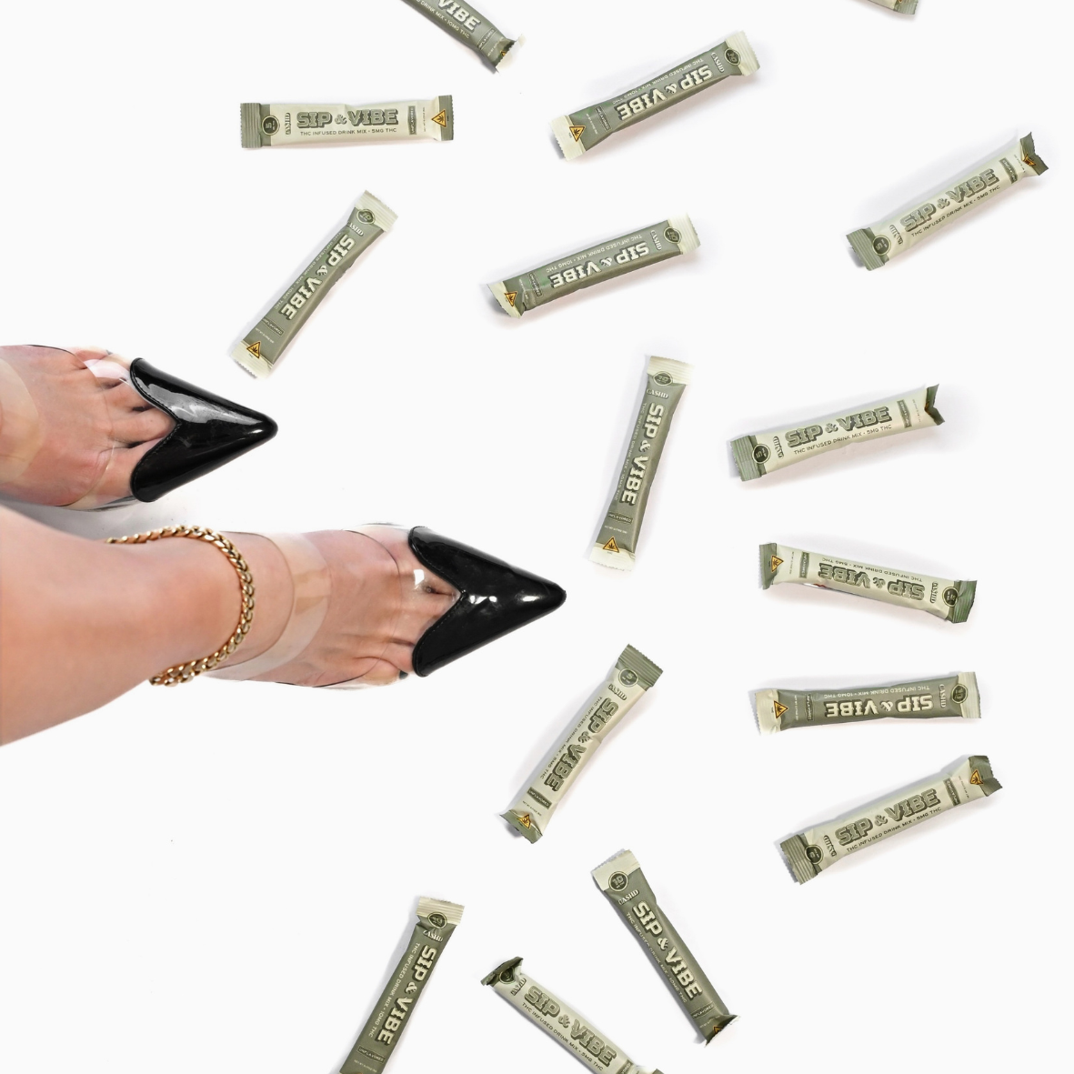 A person's feet wearing black high heels and an ankle bracelet, surrounded by scattered individually wrapped green tea flavor sachets of Sip & Vibe THC-infused drink.