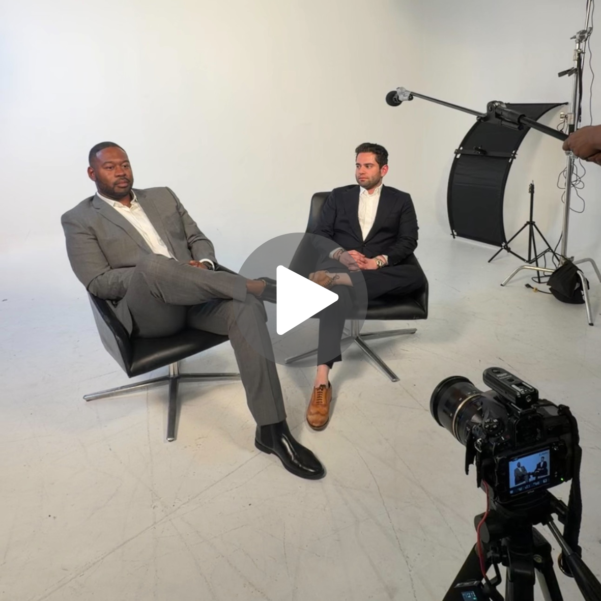 Two men in suits sitting in chairs during a studio interview or discussion, with filming equipment and a camera in a white studio space.