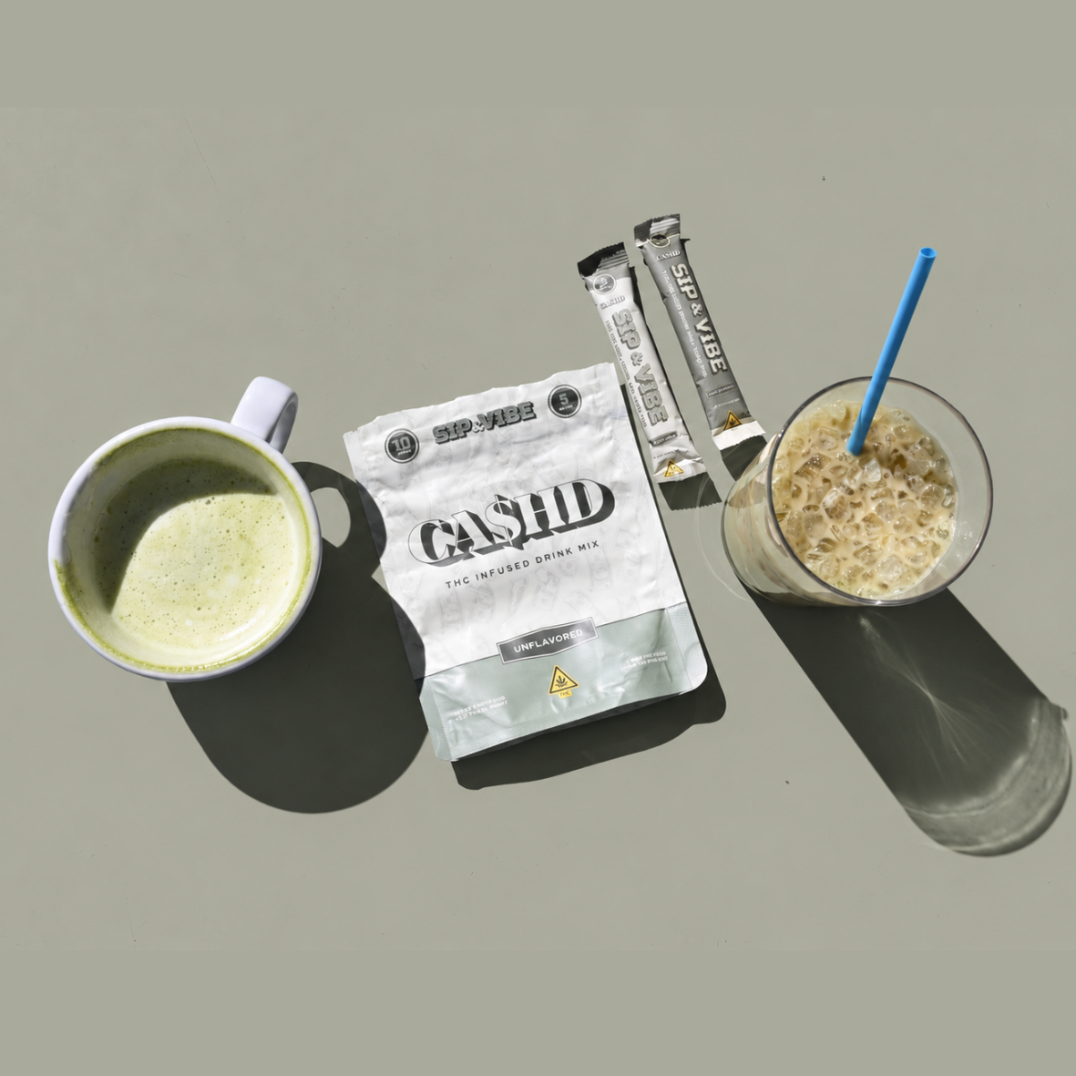 A mug of green cannabis-infused hot drink, a package of Cashid THC drink mix, two cannabis-infused bite-sized snack bars, and a glass of iced coffee with a straw on a light gray surface.
