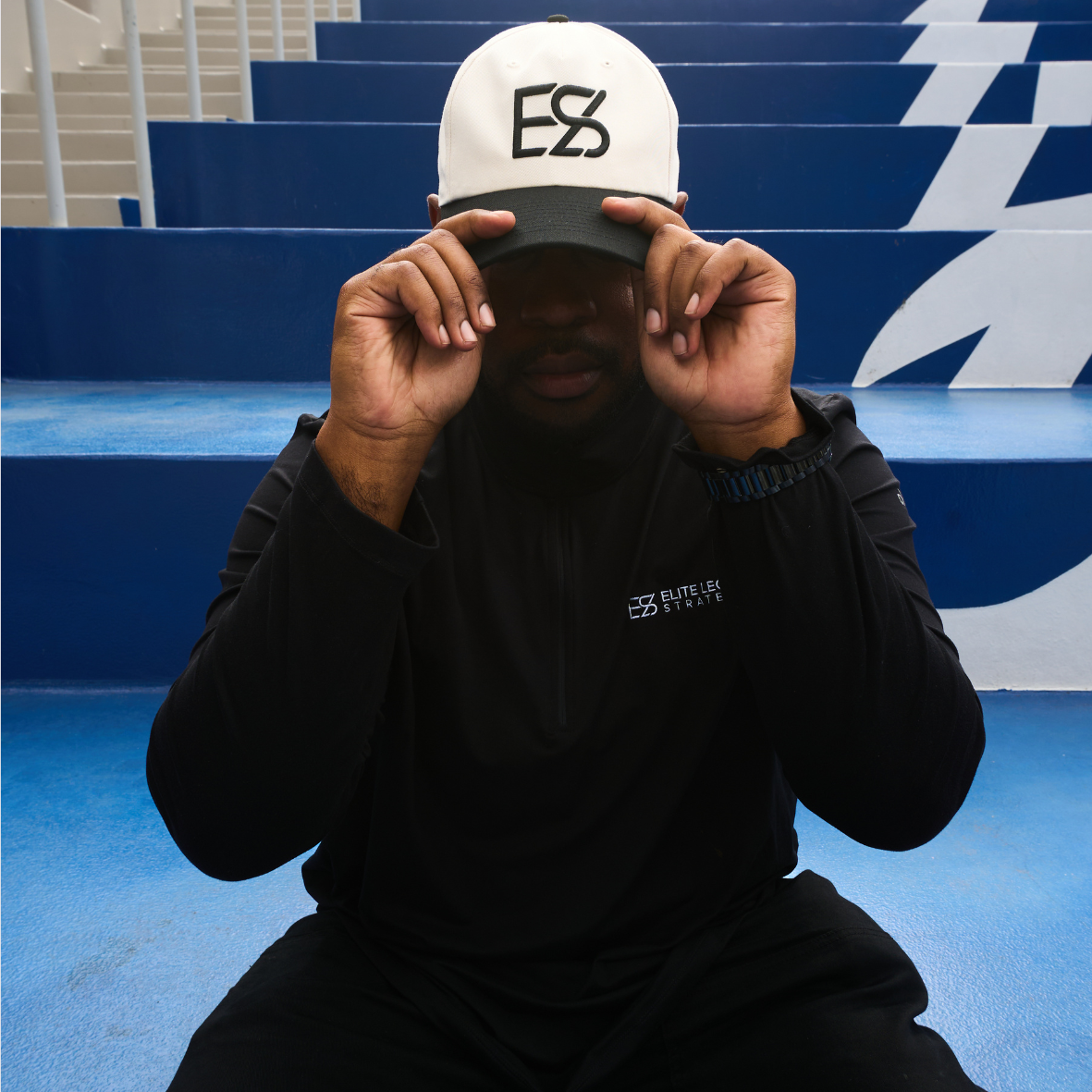 A man sitting on blue bleachers, wearing a black jacket with a logo and a white baseball cap with 'ES' on it, adjusting the cap's brim with both hands.