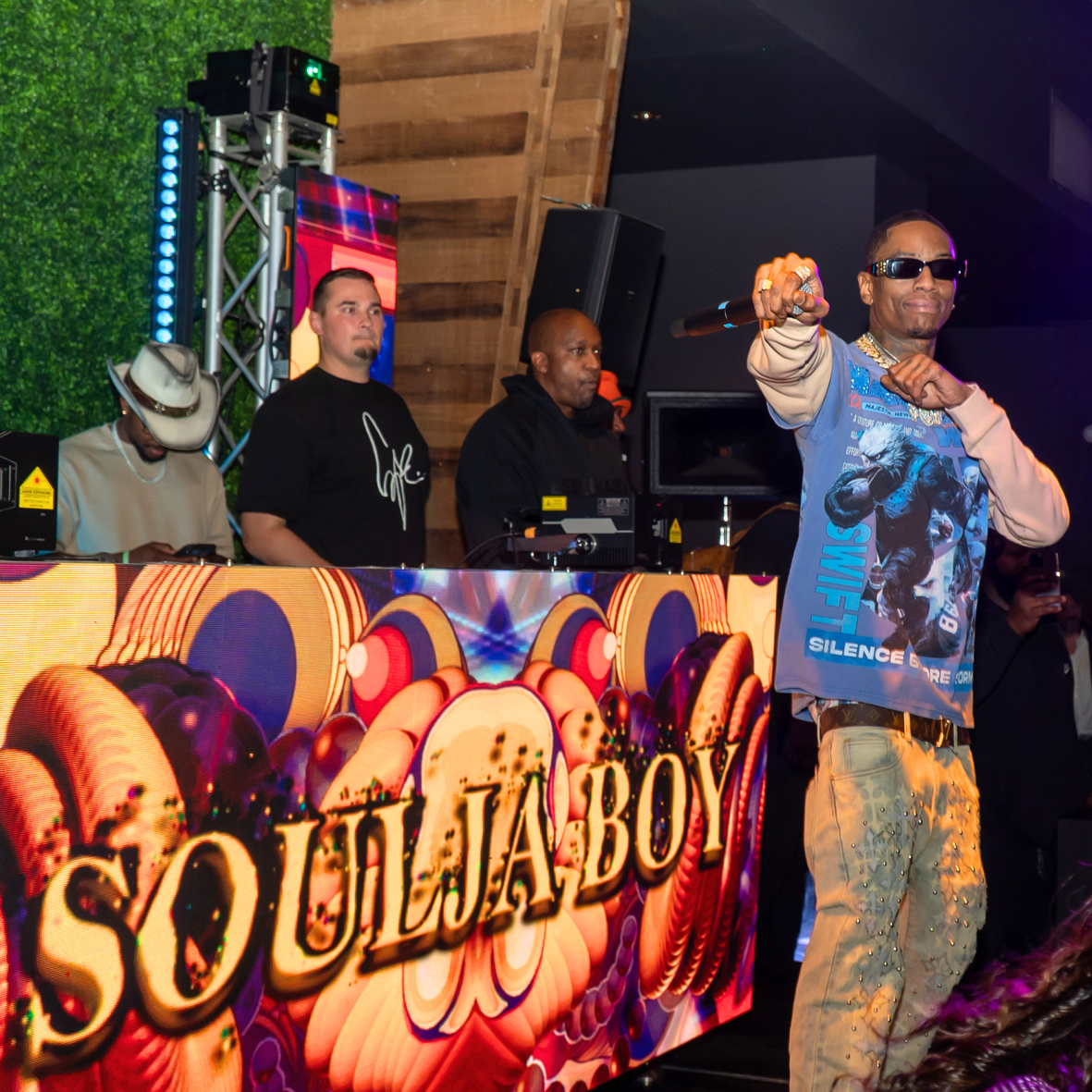 A rapper performs on stage with a DJ setup and a DJ behind him, featuring a colorful SOL LULA BOY sign. The rapper wears sunglasses, a graphic shirt, and beige pants, holding a microphone.