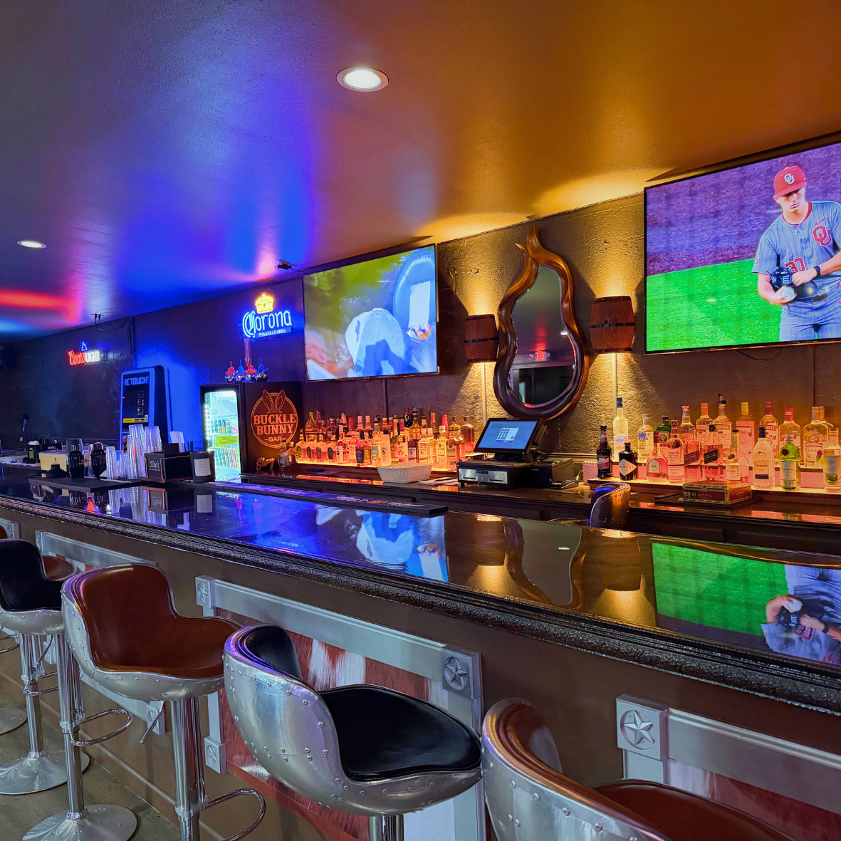 Inside a bar with a black countertop, bar stools, multiple liquor bottles behind the bar, two large TV screens showing a baseball game, neon signs including Corona and Coors Light, and a decorative mirror on the wall.