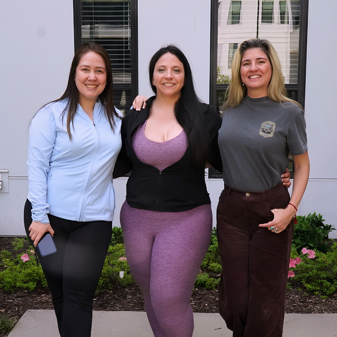 Three women standing outdoors in front of a building, smiling, with small flowers and greenery in the foreground.