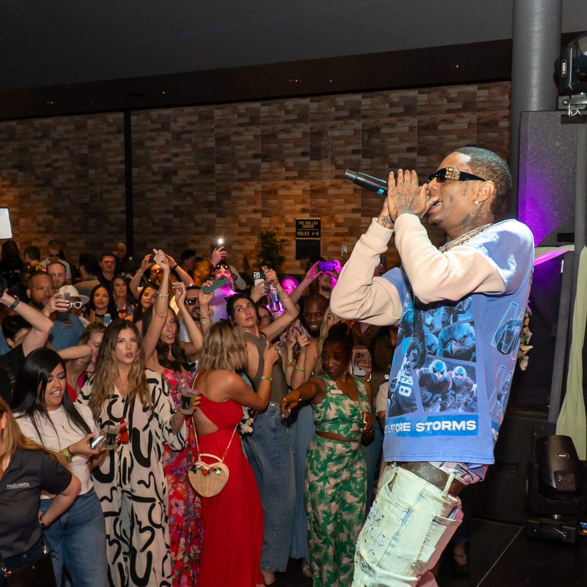 Male performer singing into a microphone on stage while a crowd of people watch and take photos at an indoor event.