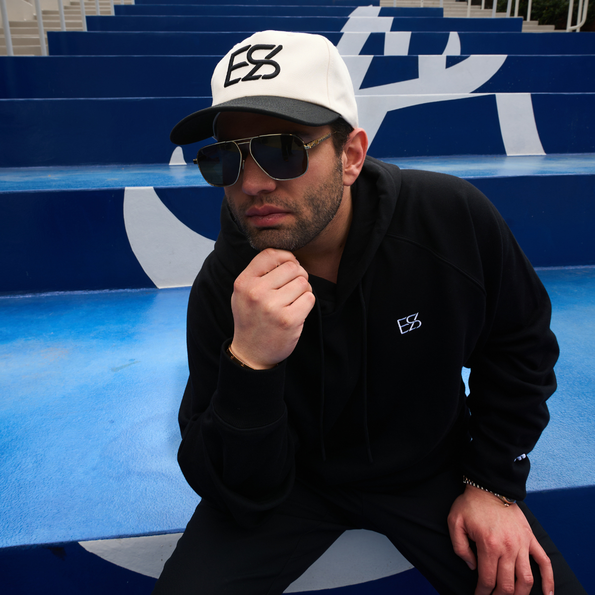 A man in sunglasses and a black hoodie sitting on blue stairs with geometric patterns, wearing a white cap with the letters 'ESS' on it.