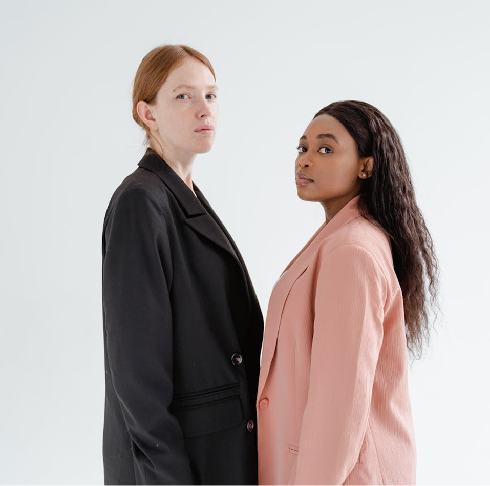 Two women standing side by side, one with red hair wearing a black blazer and the other with curly dark hair wearing a pink blazer, against a plain light background.