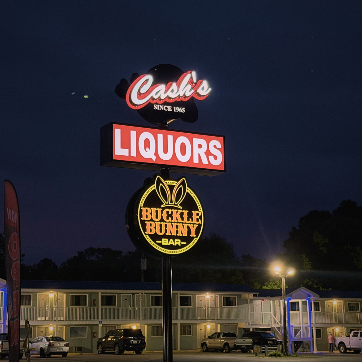 Nighttime view of neon signs for Cash's Liquors and Buckle Bunny Bar outside a motel with parked cars and a building with rooms.