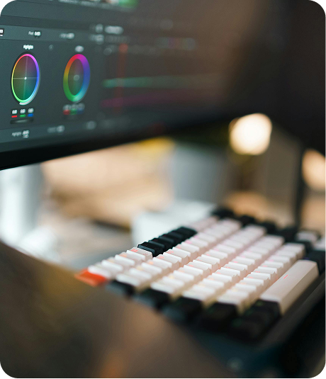 Close-up of a computer keyboard with a digital audio workstation and color wheel on the monitor in the background.