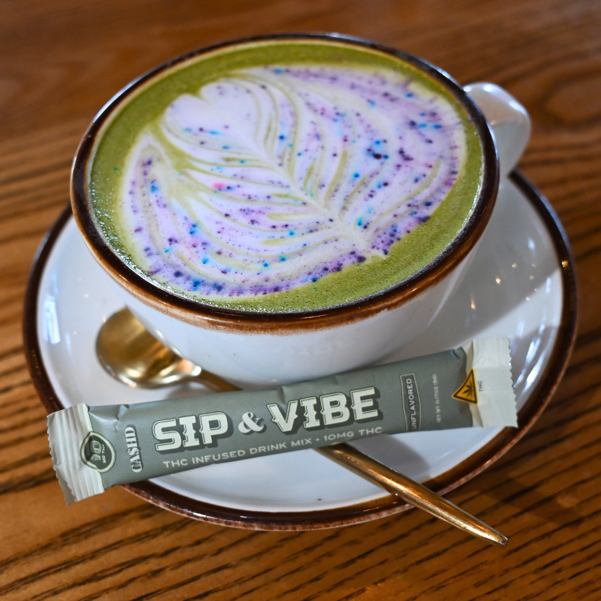 A cup of matcha latte with colorful sprinkles and latte art on top, placed on a saucer. A THC-infused drink mix packet labeled 'Sip & Vibe' is on the saucer along with a small spoon.