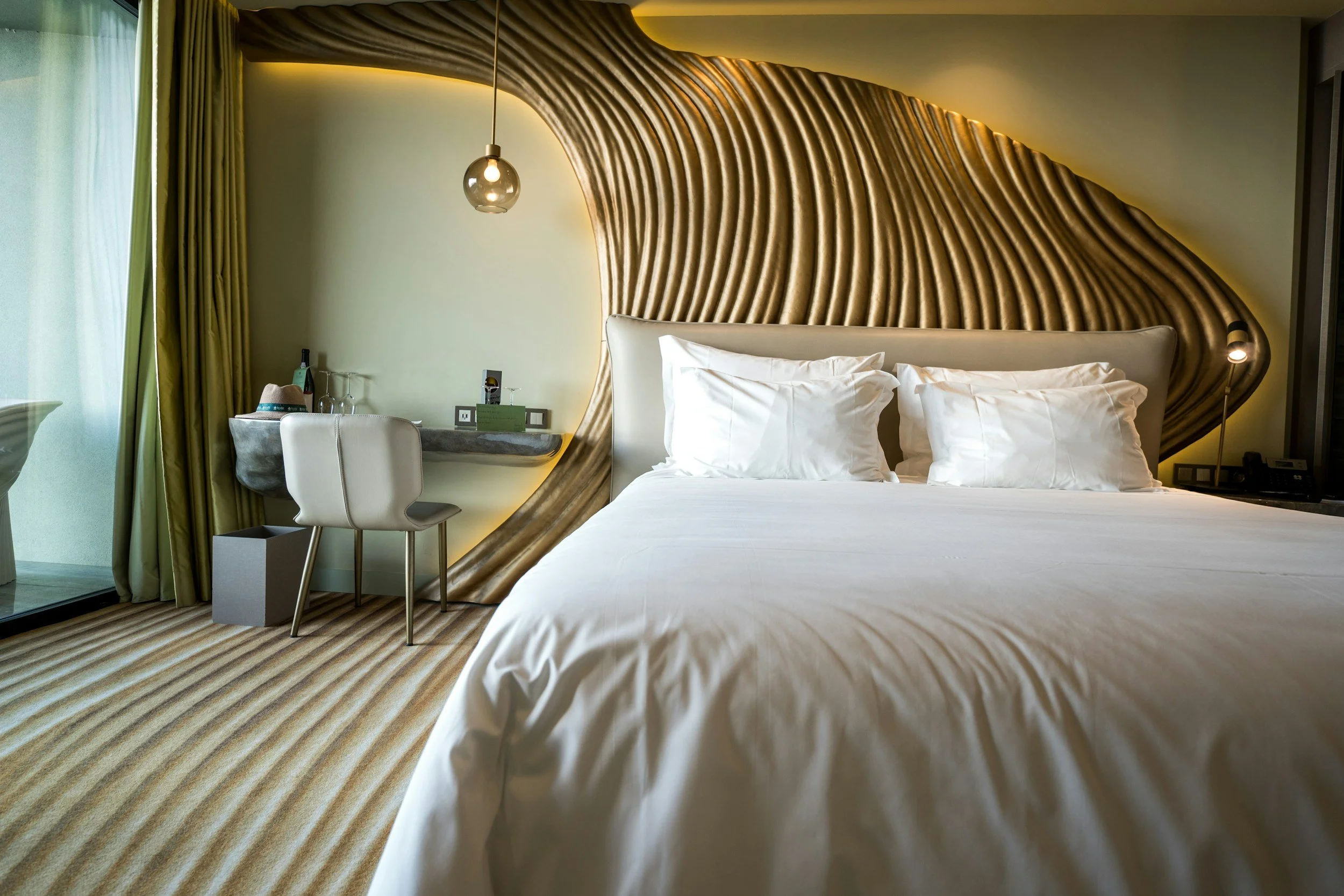 Modern hotel room with a decorative curved wooden headboard, white bed with pillows, bedside lights, a desk with a chair, and a seating area near a large window with green curtains.