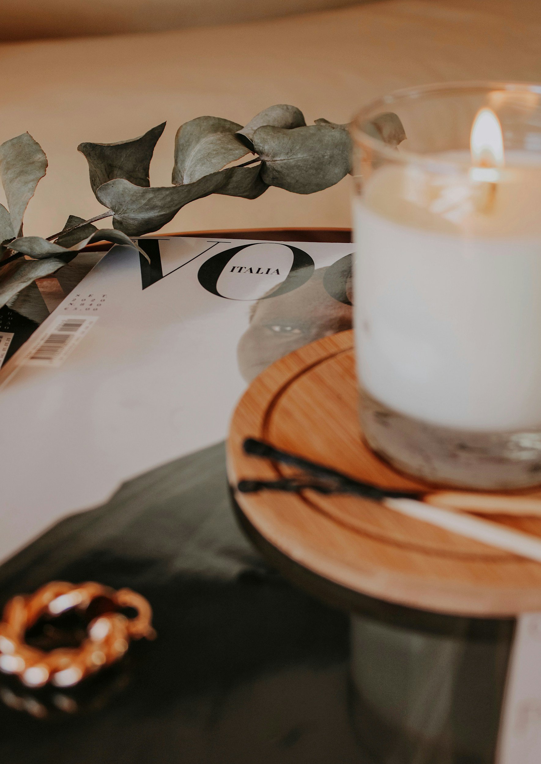 A lit white candle in a glass holder on a wooden tray, with dried eucalyptus leaves nearby, and a magazine with the word 'Italia' on the cover.