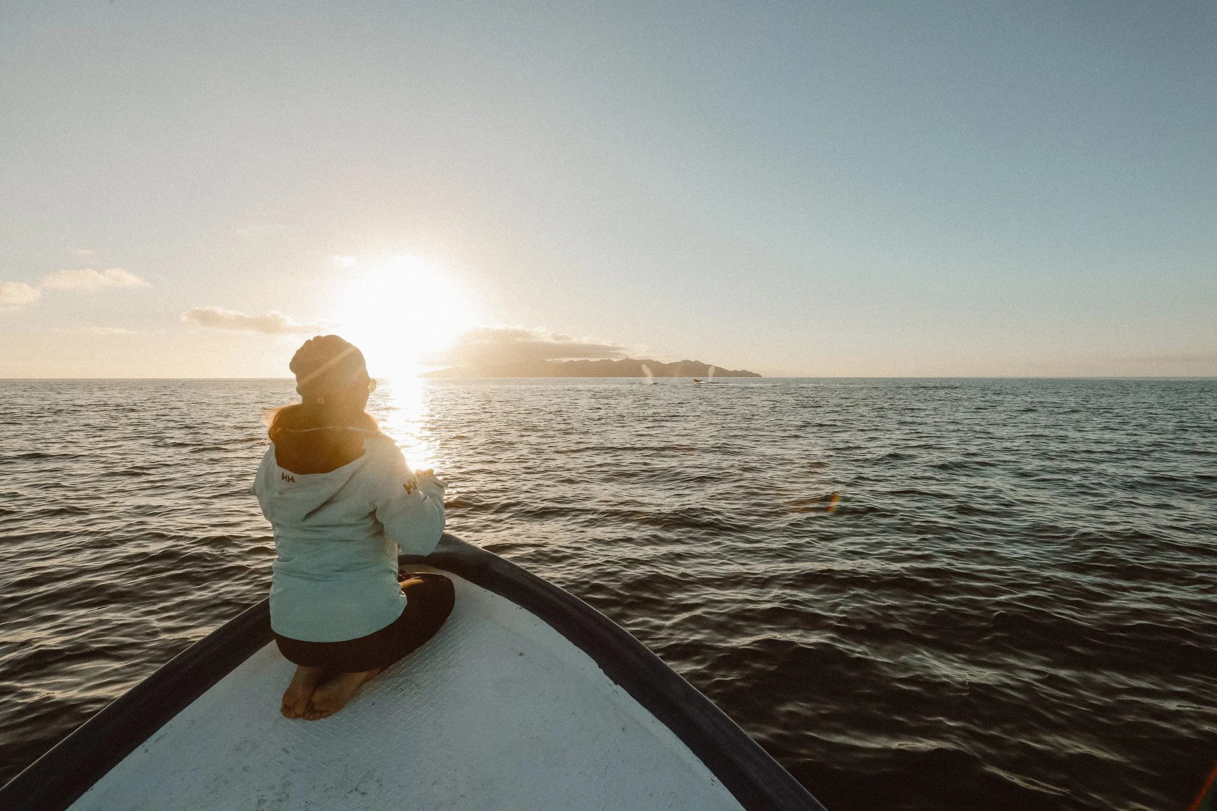 A person sitting on a surfboard at the ocean during sunset, facing away, with mountains and boats in the distance.