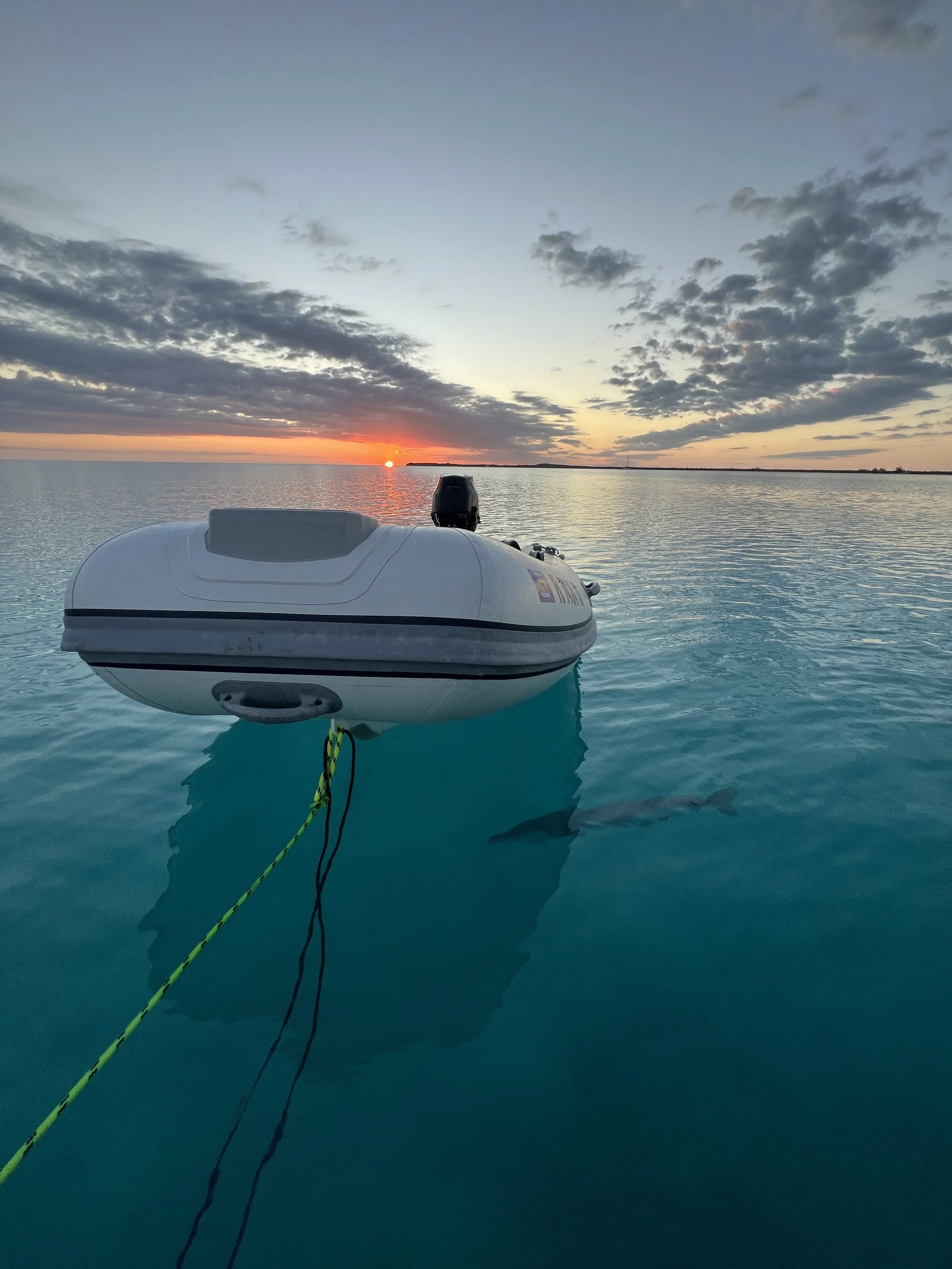 A small inflatable boat with an outboard motor on calm water during sunset, with a colorful sky and scattered clouds in the background.