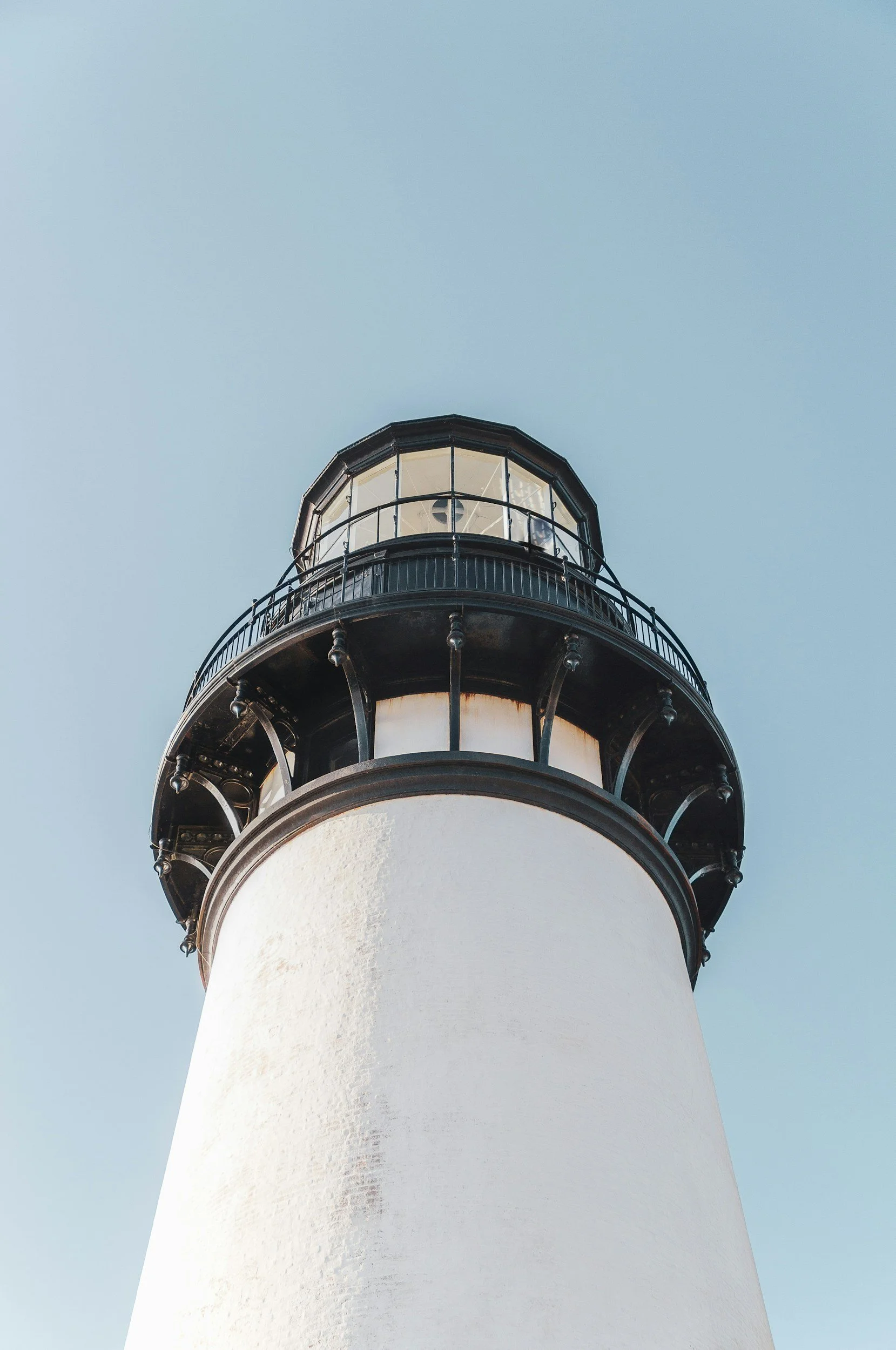 A tall white lighthouse with black metal balcony railings near the top, set against a clear blue sky.