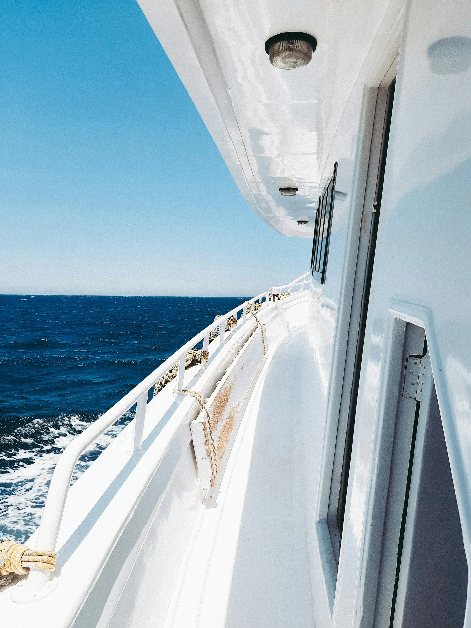 View of the side of a white ship with a blue sky and open ocean in the background, showing a rails, port lights, and part of the ship's cabin.