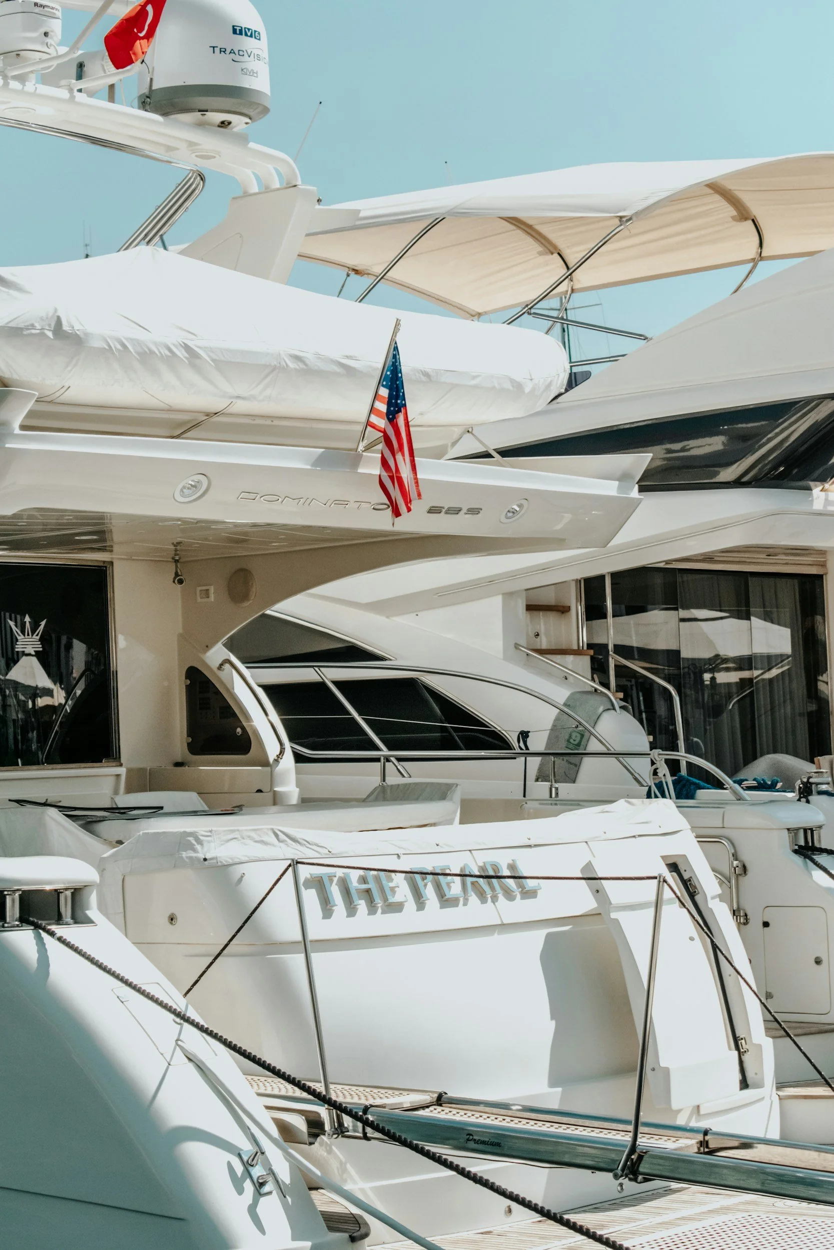 Multiple luxury yachts docked at a marina, visible with white exteriors, some with umbrellas and awnings, and American flags on one yacht
