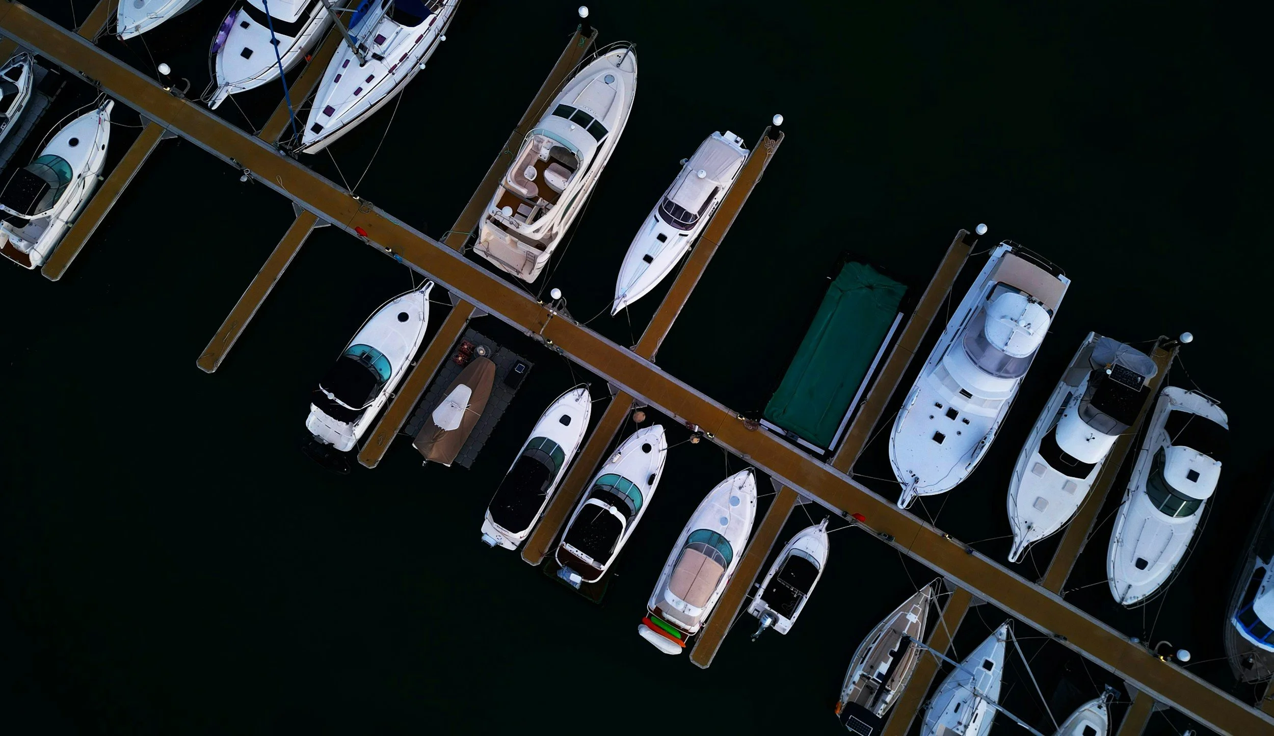 An aerial view of boats docked at a marina, arranged in parallel rows along floating docks on calm water.