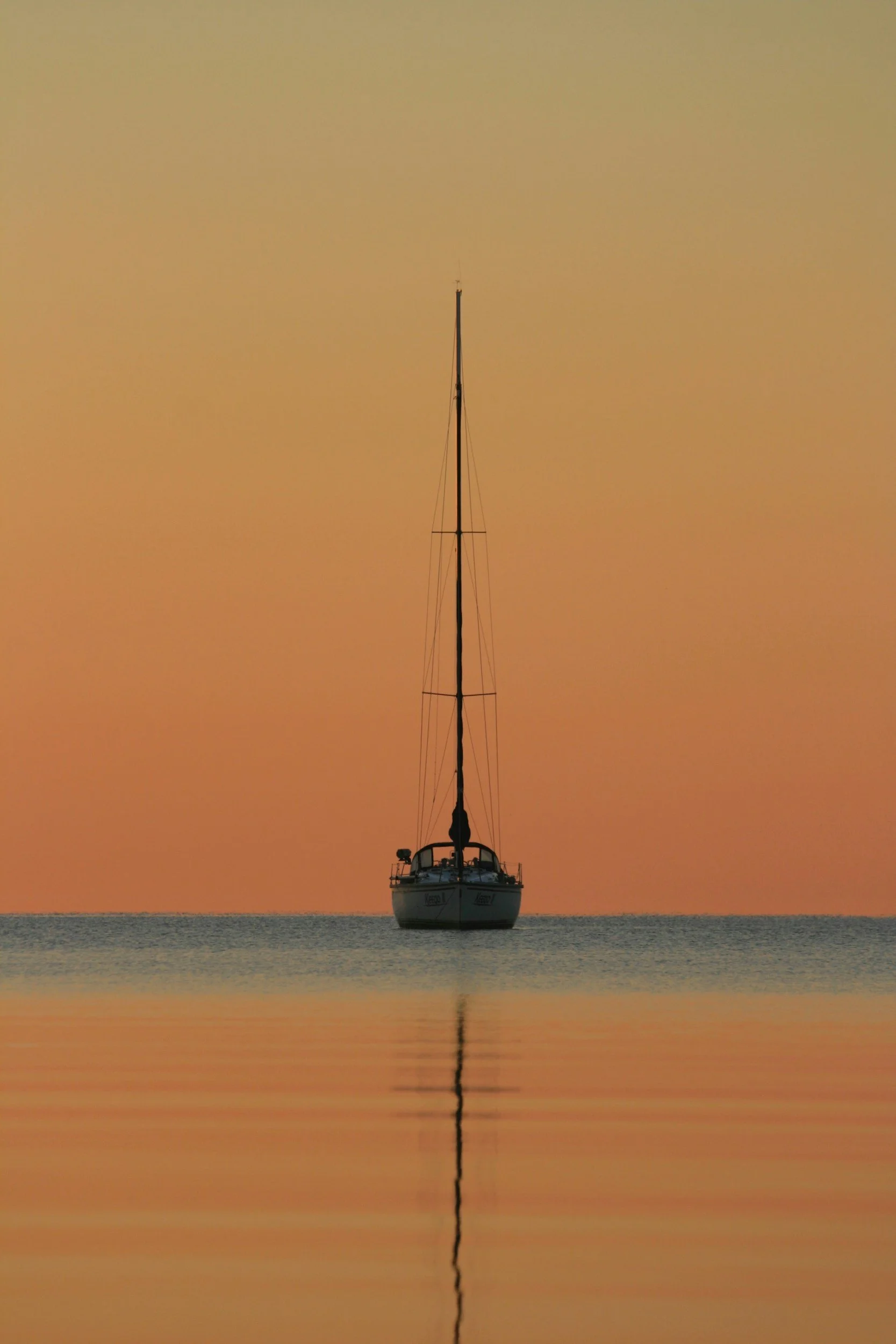 A sailboat floating on calm water during sunset with a colorful orange sky and no clouds.