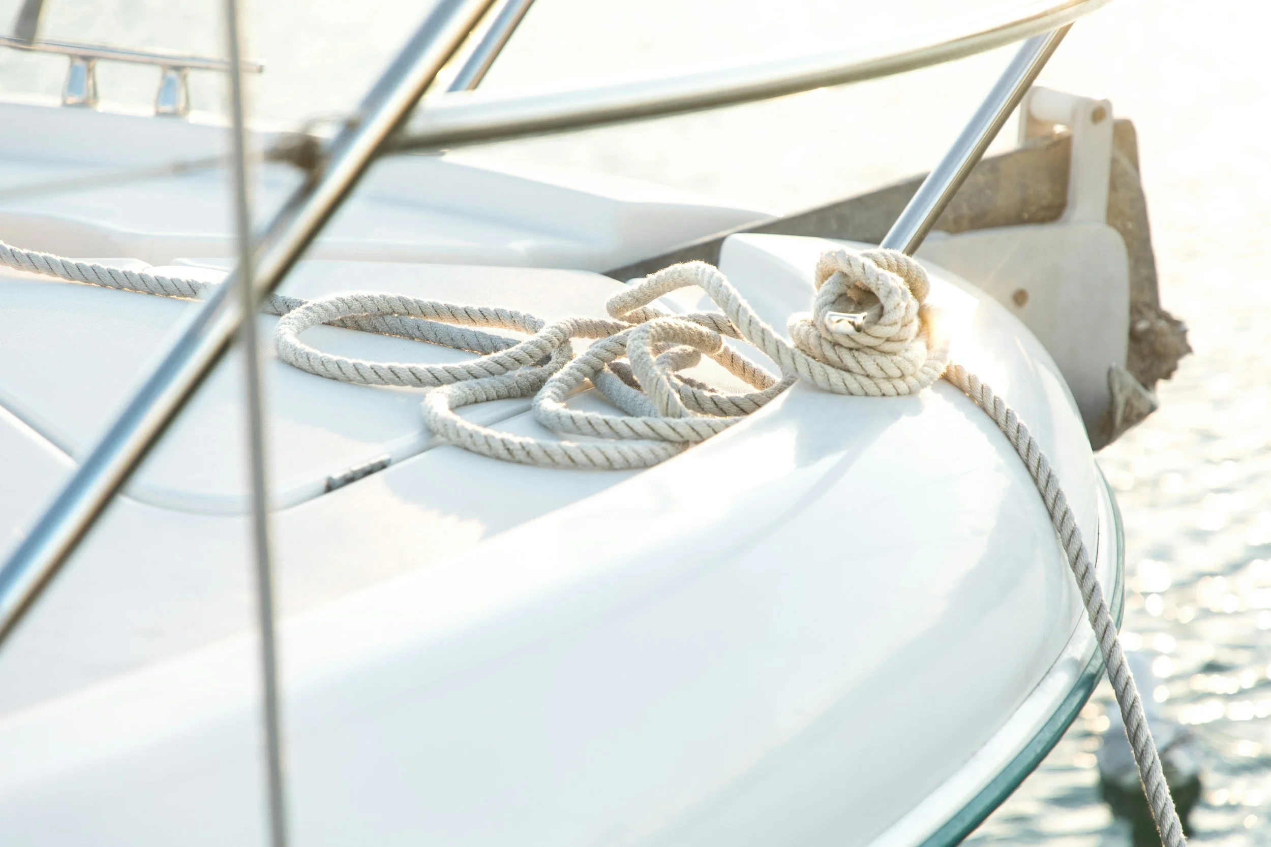Close-up of a white boat with thick rope tied around the deck, secured with a metal cleat, on calm water during daylight.