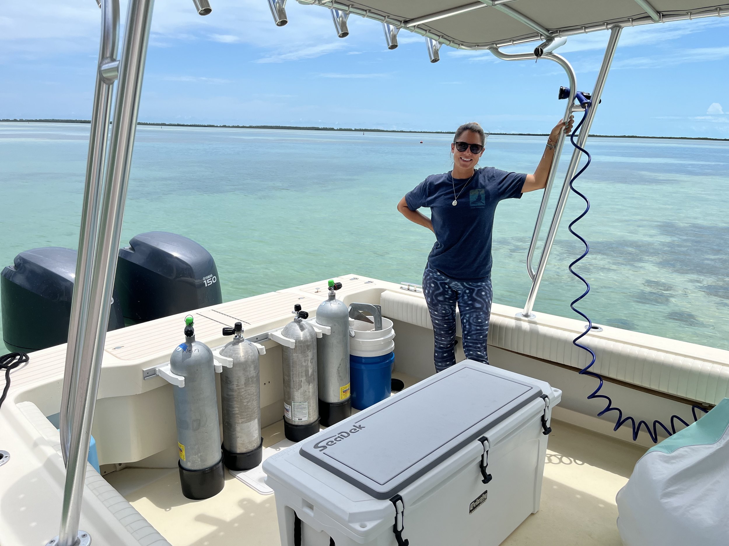 A woman standing on a boat holding a metal rail above her head with a body of water and a cloudy sky in the background. The boat has various equipment, including oxygen tanks, a white cooler, and other gear, indicating it is used for diving or marine