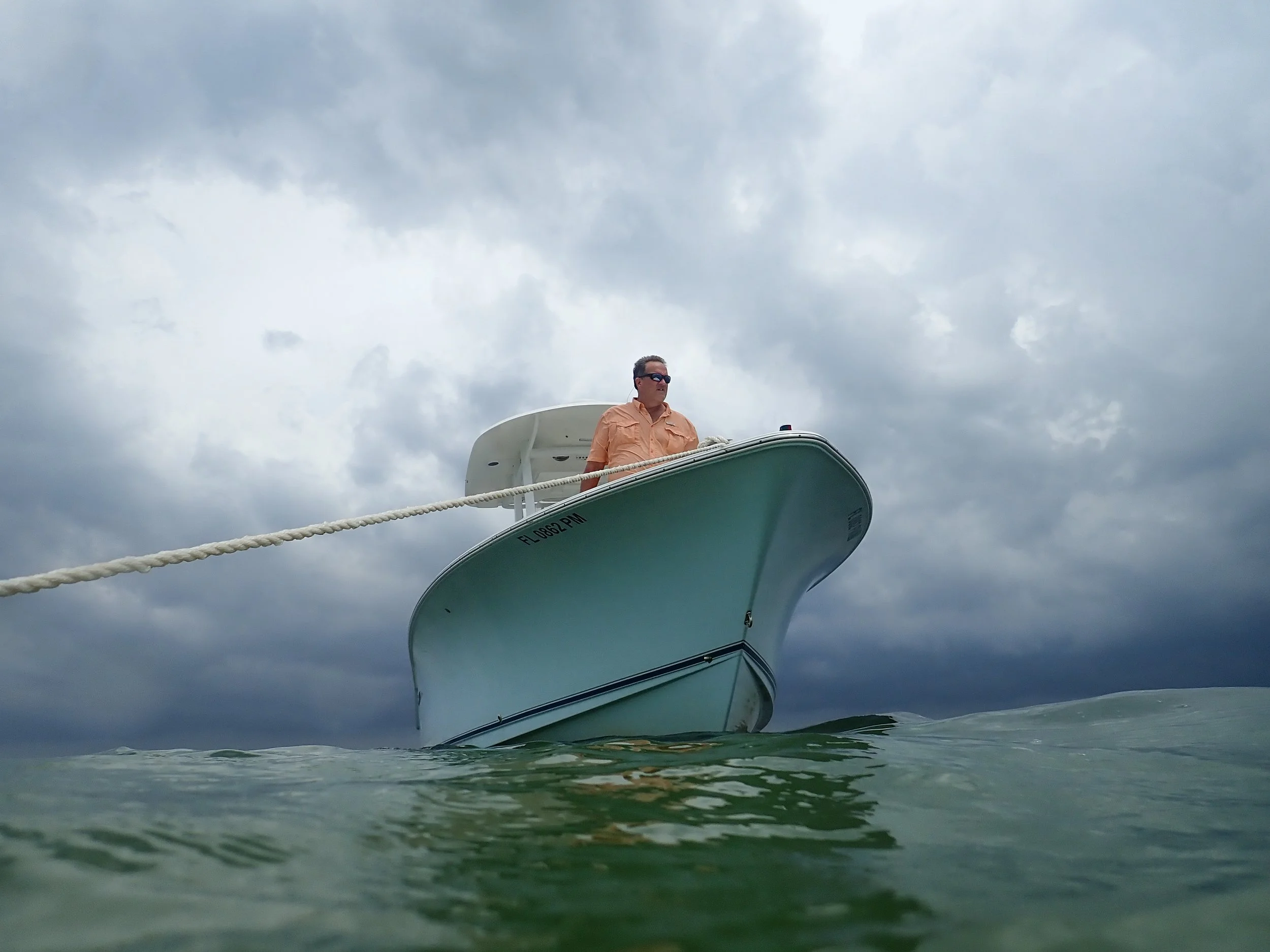 Man in sunglasses standing on the bow of a boat during overcast weather, with water and cloudy sky in the background.