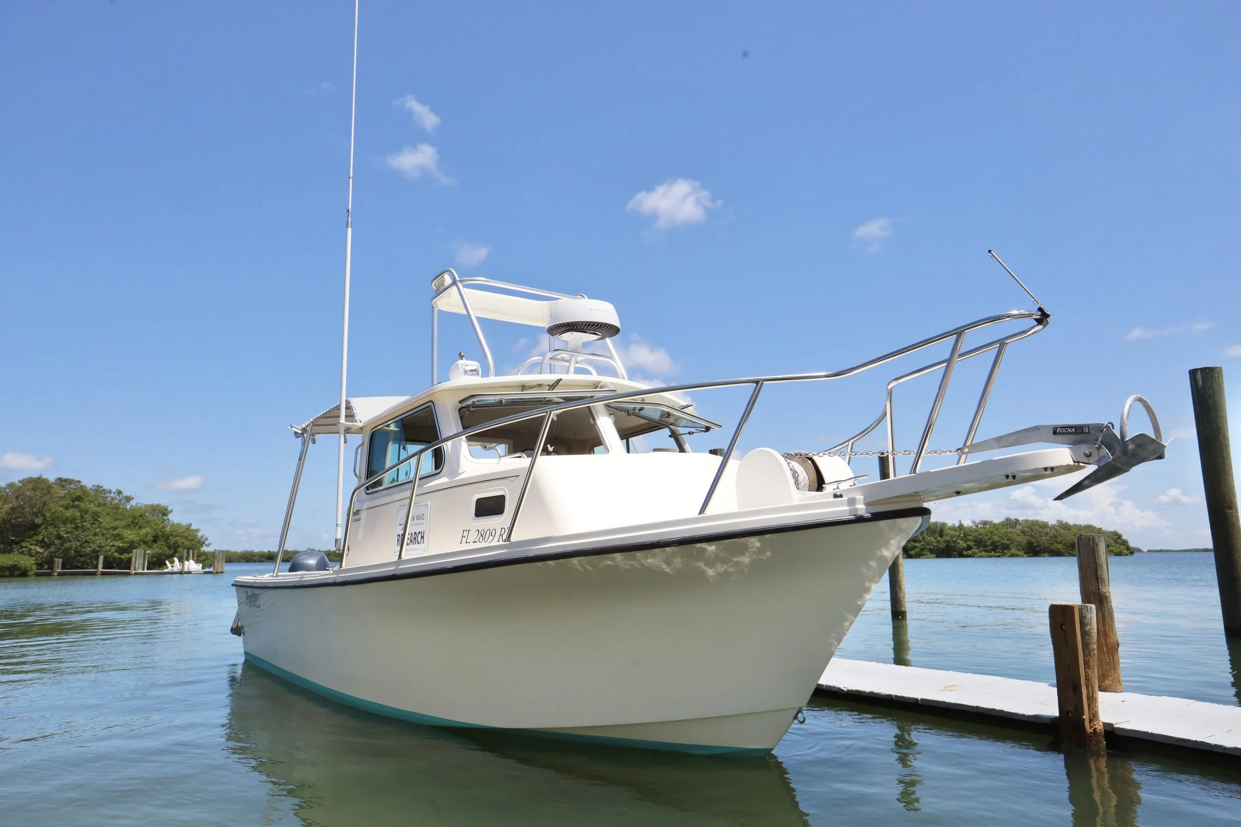 A white boat docked at a pier on calm water with green trees and a blue sky with a few clouds in the background.