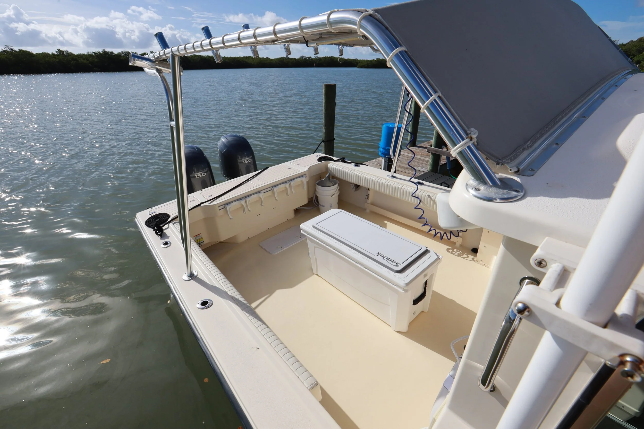 A boat docked on the water with a gray canopy, two outboard engines, and various equipment on the deck, with trees and a cloudy sky in the background.