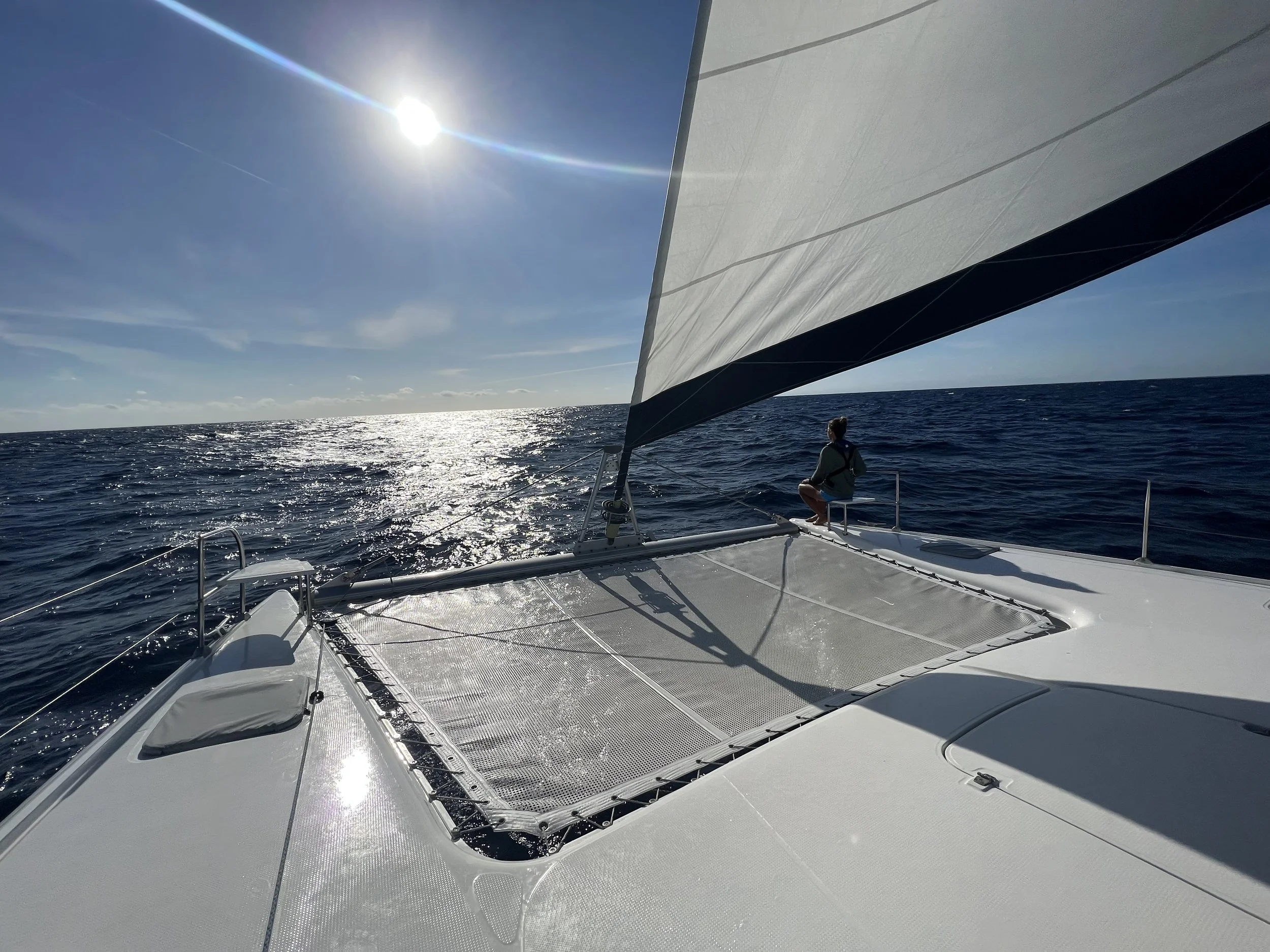 A person sitting on the deck of a sailboat in the middle of open ocean under a clear sky, with the sun shining brightly.