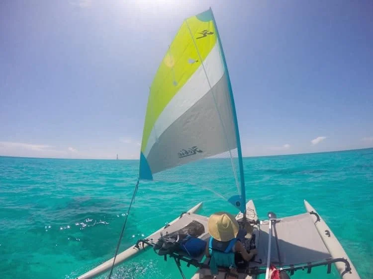 Person sailing a small boat with a colorful sail on clear turquoise water under a bright sunny sky.