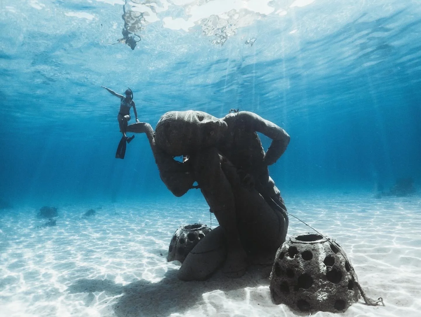 An underwater scene featuring a diver with fins near a large sculpture of a reclining man made of rock, with two hollowed rocks at the base, and a snorkeler swimming above the sculpture, in clear blue water.
