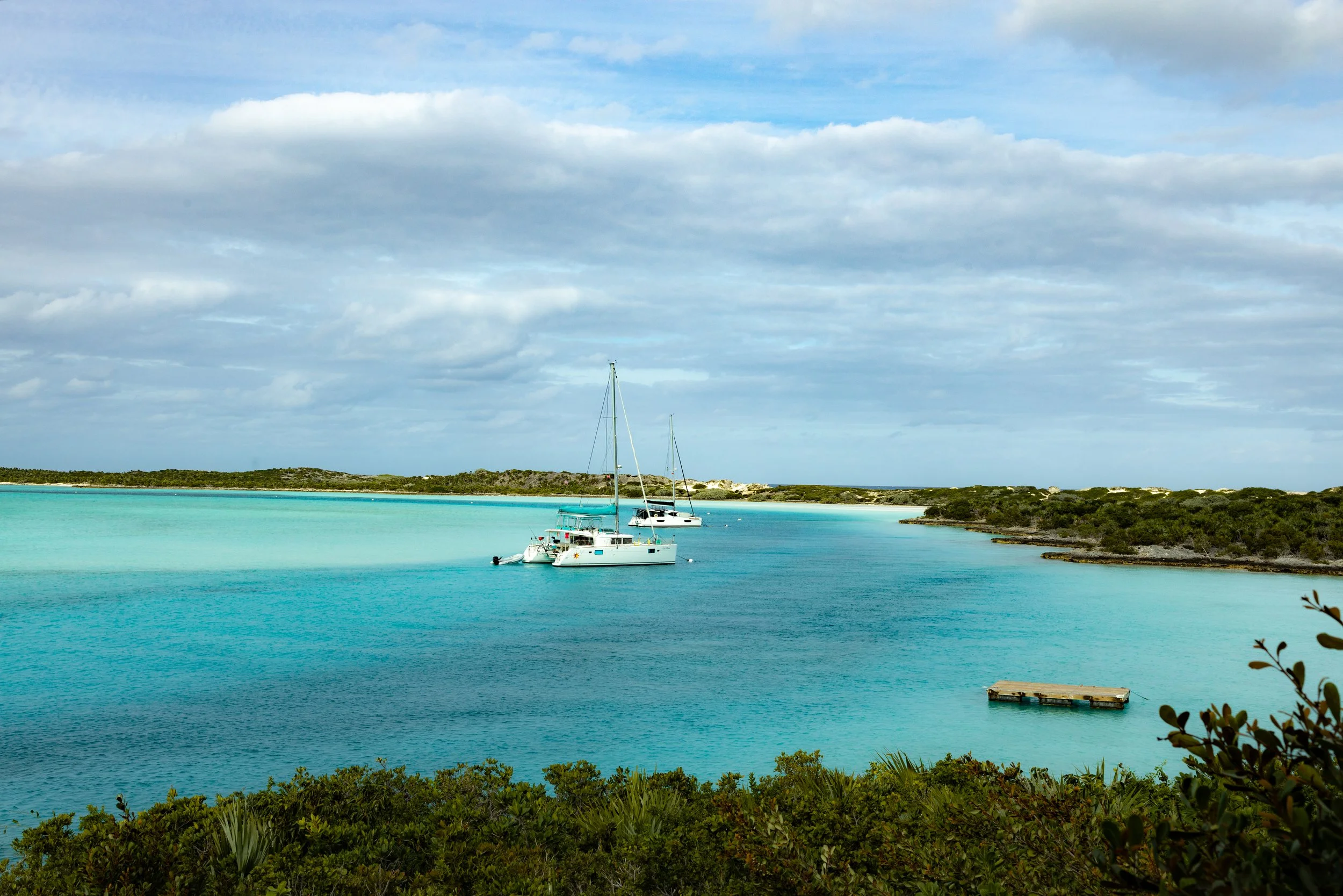 A scenic view of a turquoise bay with a sailboat and a motorboat anchored near the shore. The background features a cloudy sky and a distant strip of land with low vegetation and small trees. Foreground includes lush green shrubs and plants.
