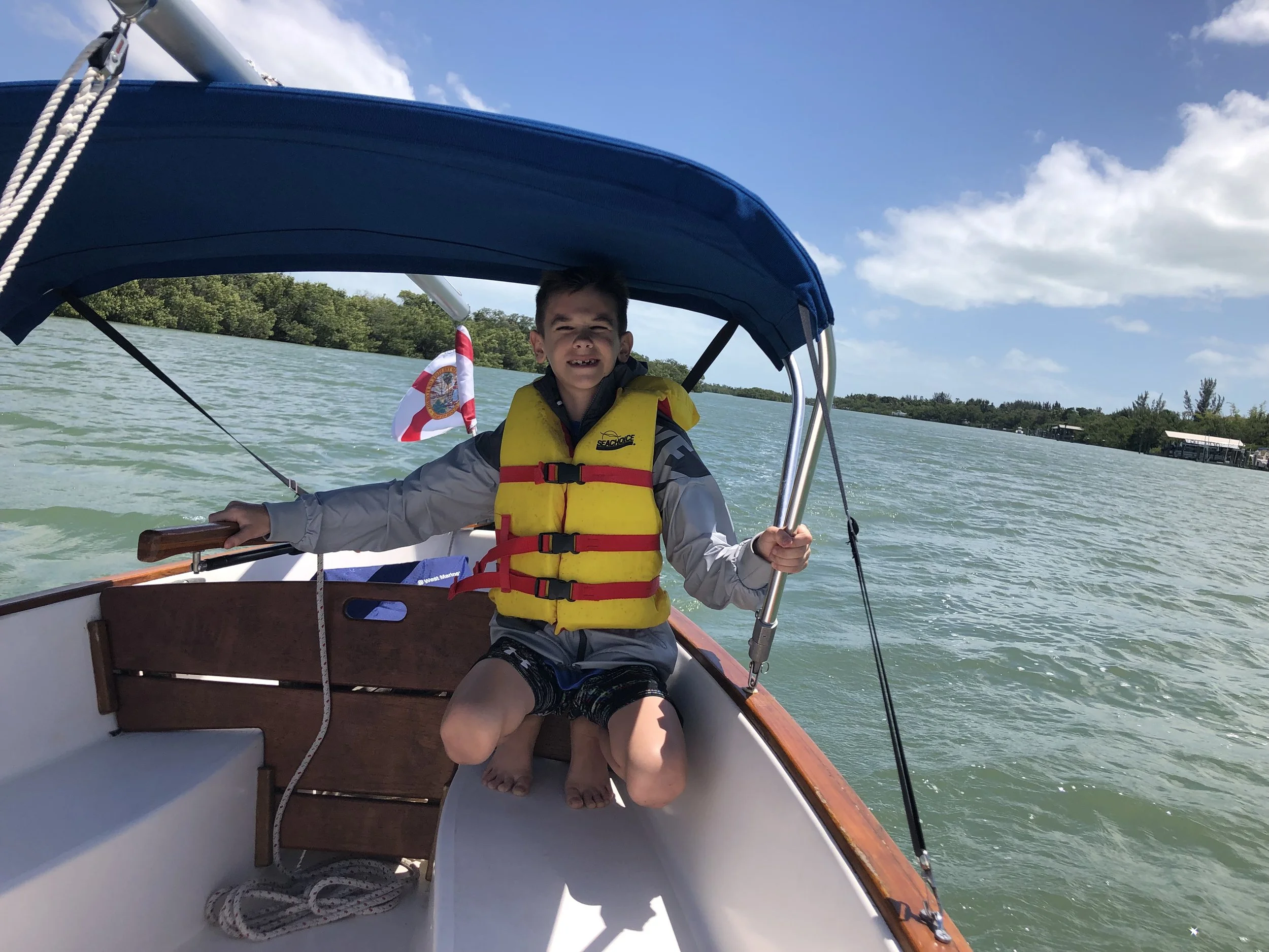 A young boy sitting on a boat, wearing a yellow life jacket, caught smiling with missing teeth, enjoying a boat ride on a clear day with a blue sky, water, and trees in the background.