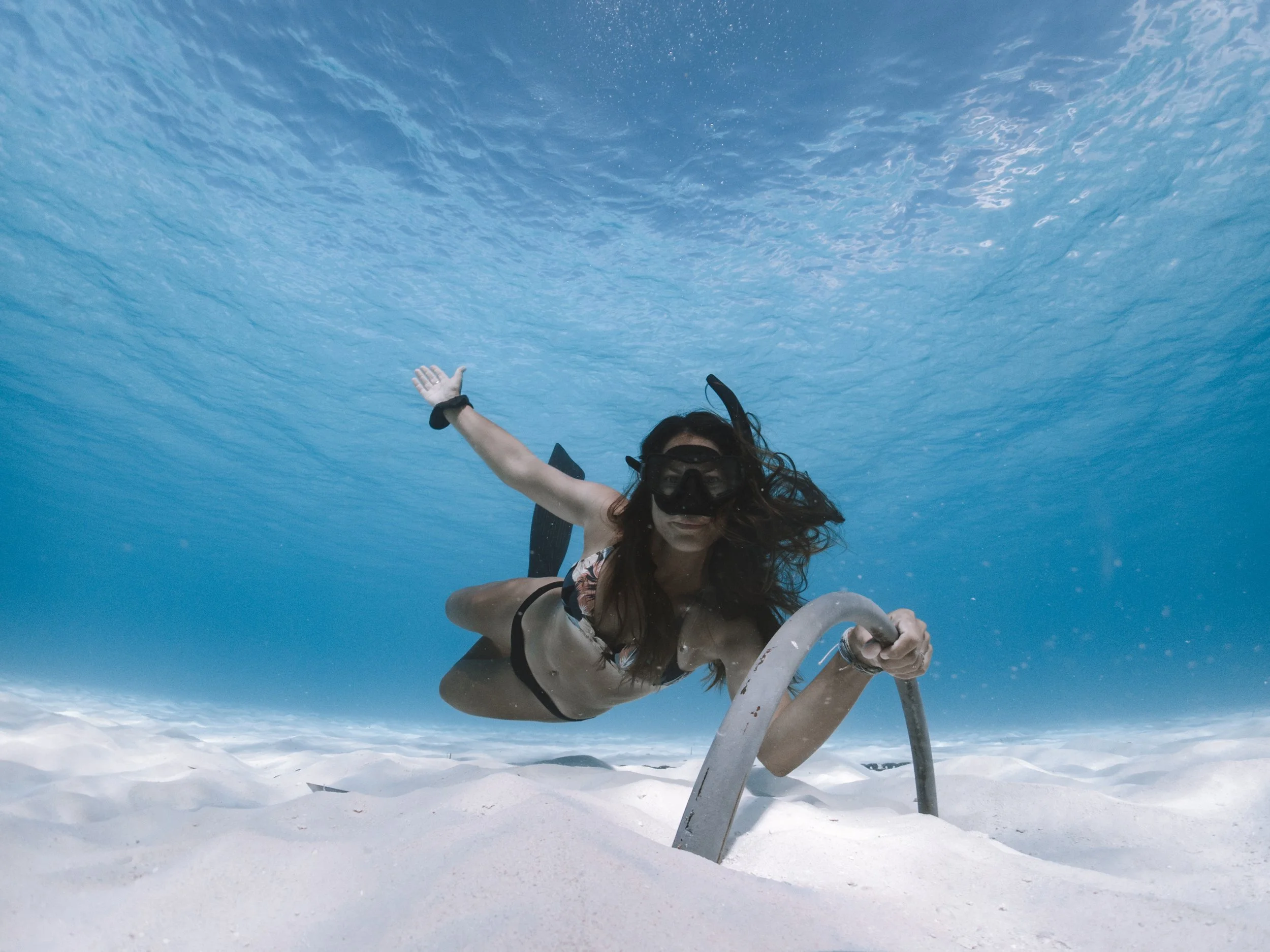 Woman in swimming goggles and swimsuit underwater holding a sun lounger.