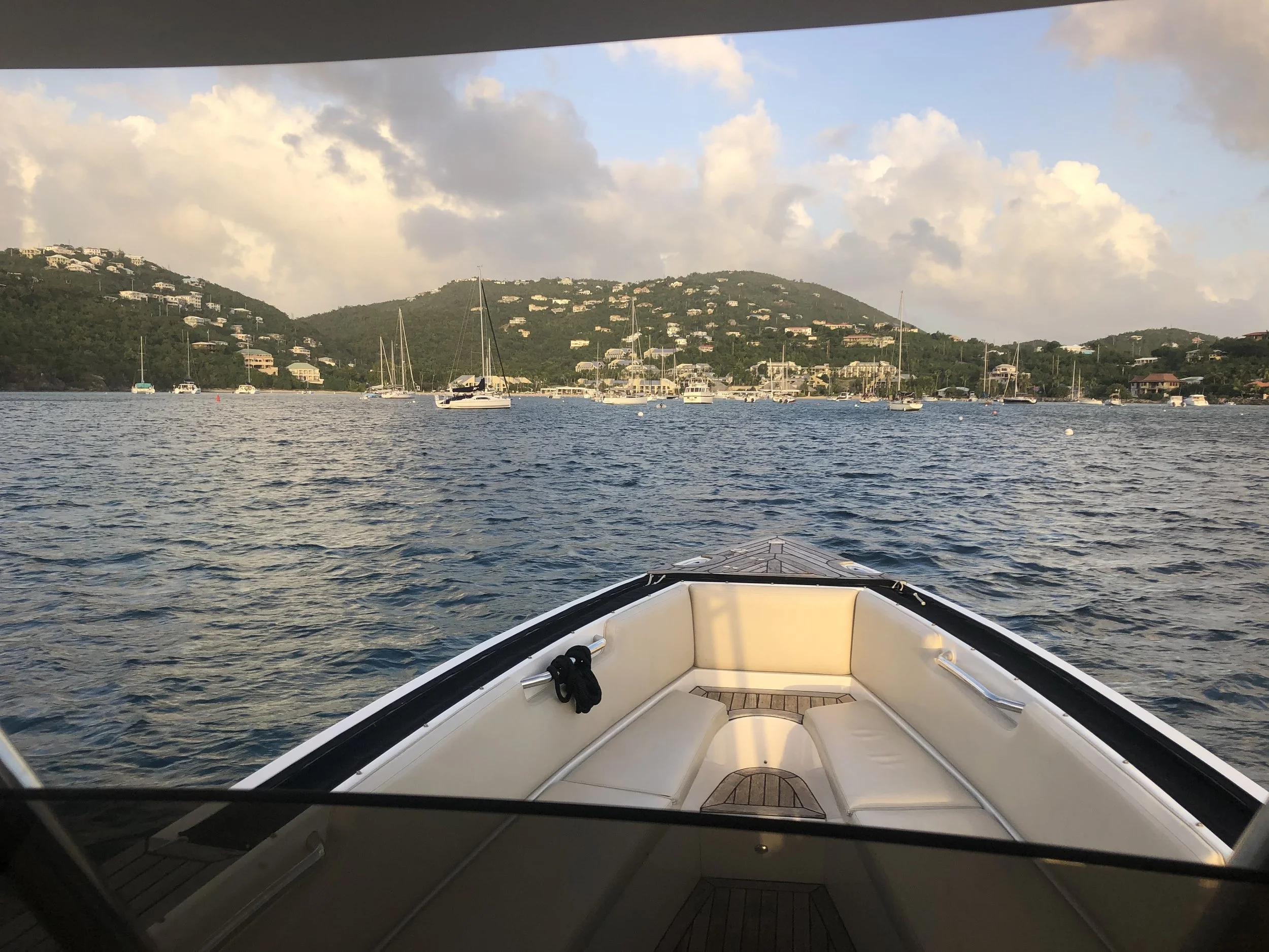 View from the bow of a boat overlooking sailboats anchored in a bay with green, hilly islands and houses in the background under a partly cloudy sky.