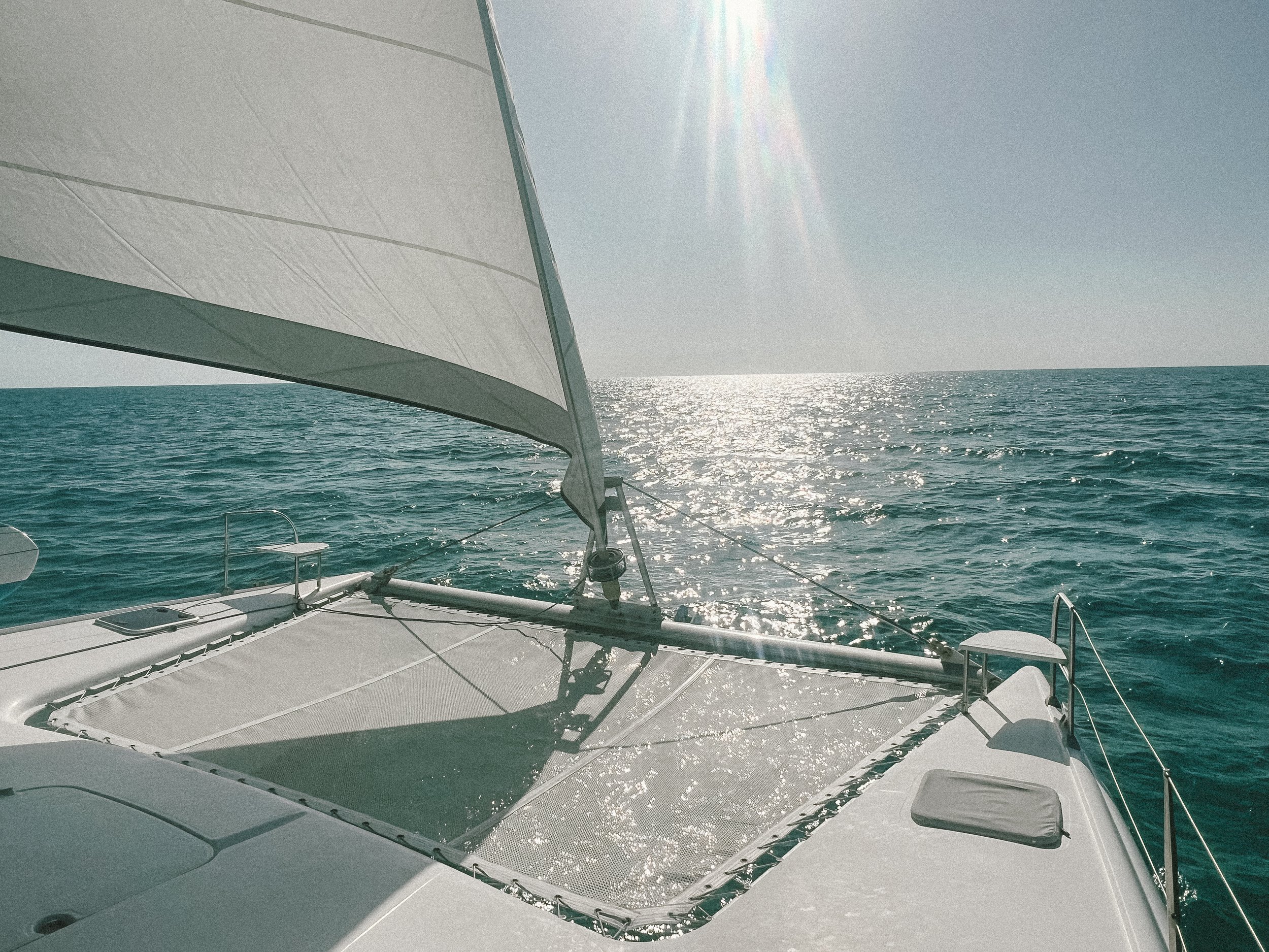 View of a sailboat deck with large white sail catching the sunlight over the ocean, horizon visible in the background.