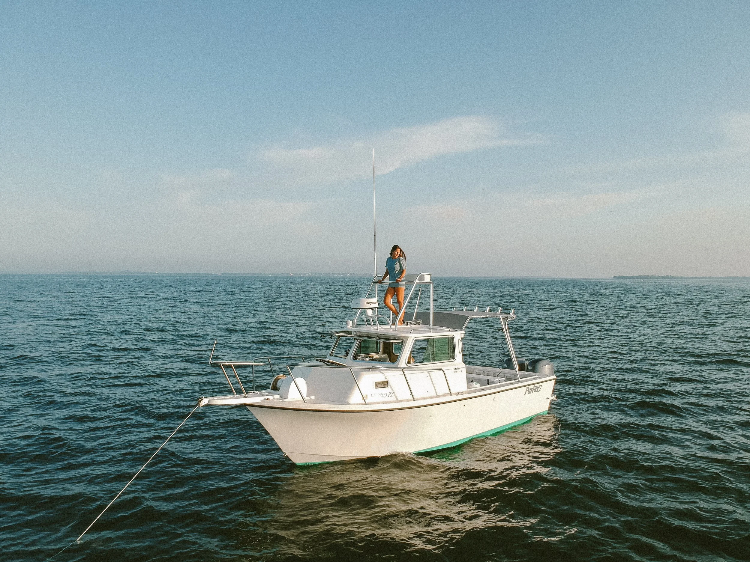A woman in a casual outfit standing on the deck of a white motorboat on calm waters, with a clear sky above.