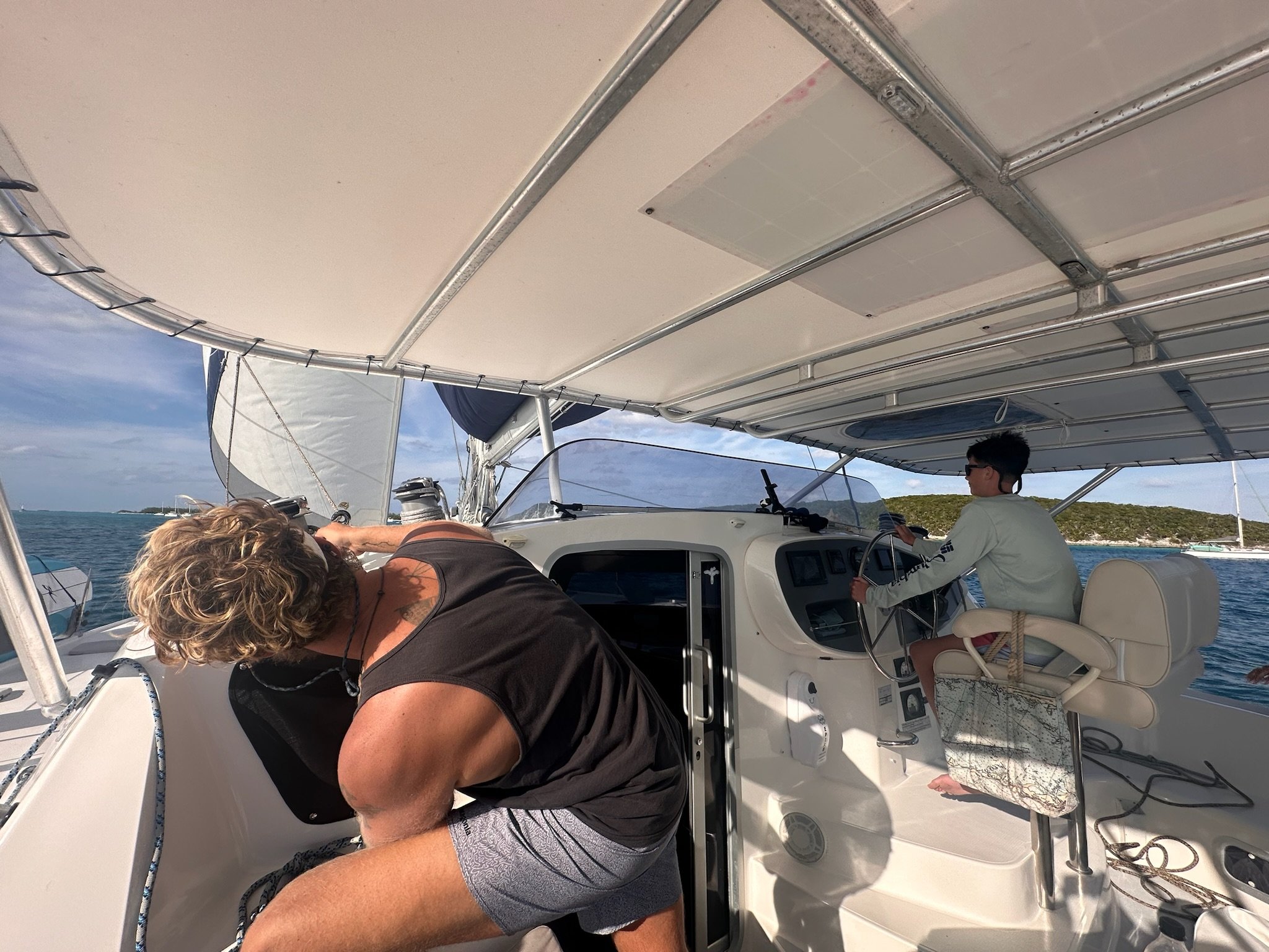 People on a boat, with one person leaning over the side and another steering, under a canopy during daytime.