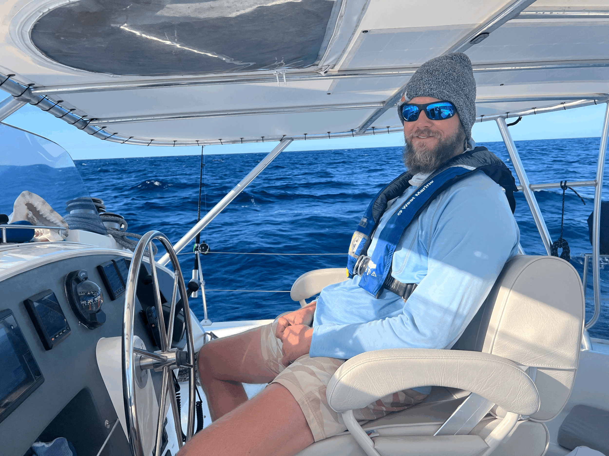 A man with a beard, wearing a gray beanie, blue reflective sunglasses, and a light blue long-sleeve shirt, is sitting on a boat in the ocean. The boat's dashboard and steering wheel are visible, and the surrounding water is deep blue with gentle wave