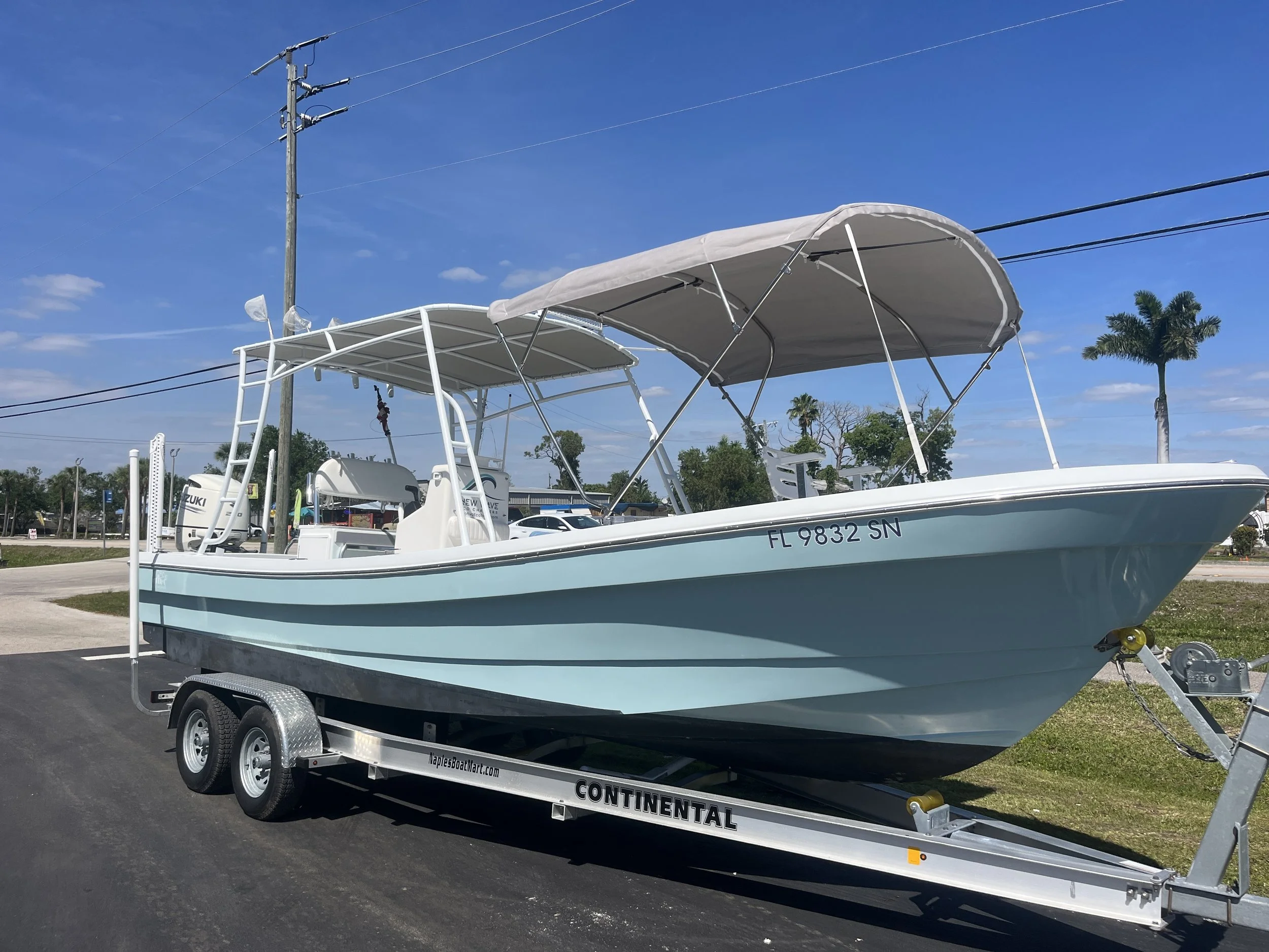 A light blue and white boat on a trailer parked on a paved area with palm trees and power lines in the background.