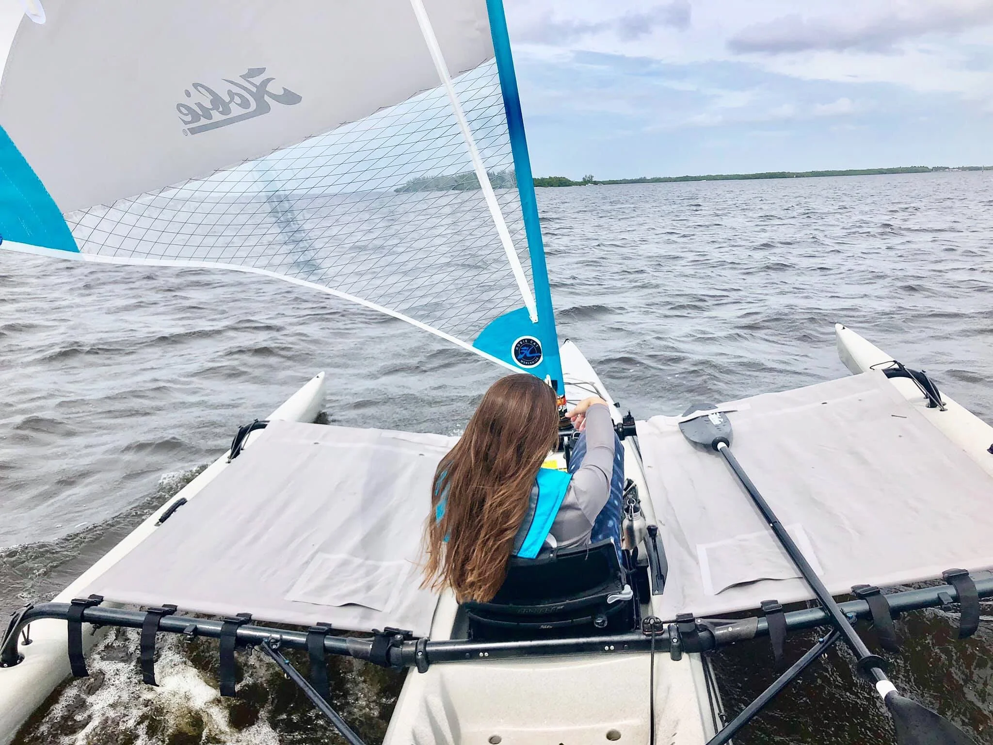 A person with long brown hair sailing a small boat on a large body of water with a distant shoreline and partly cloudy sky.