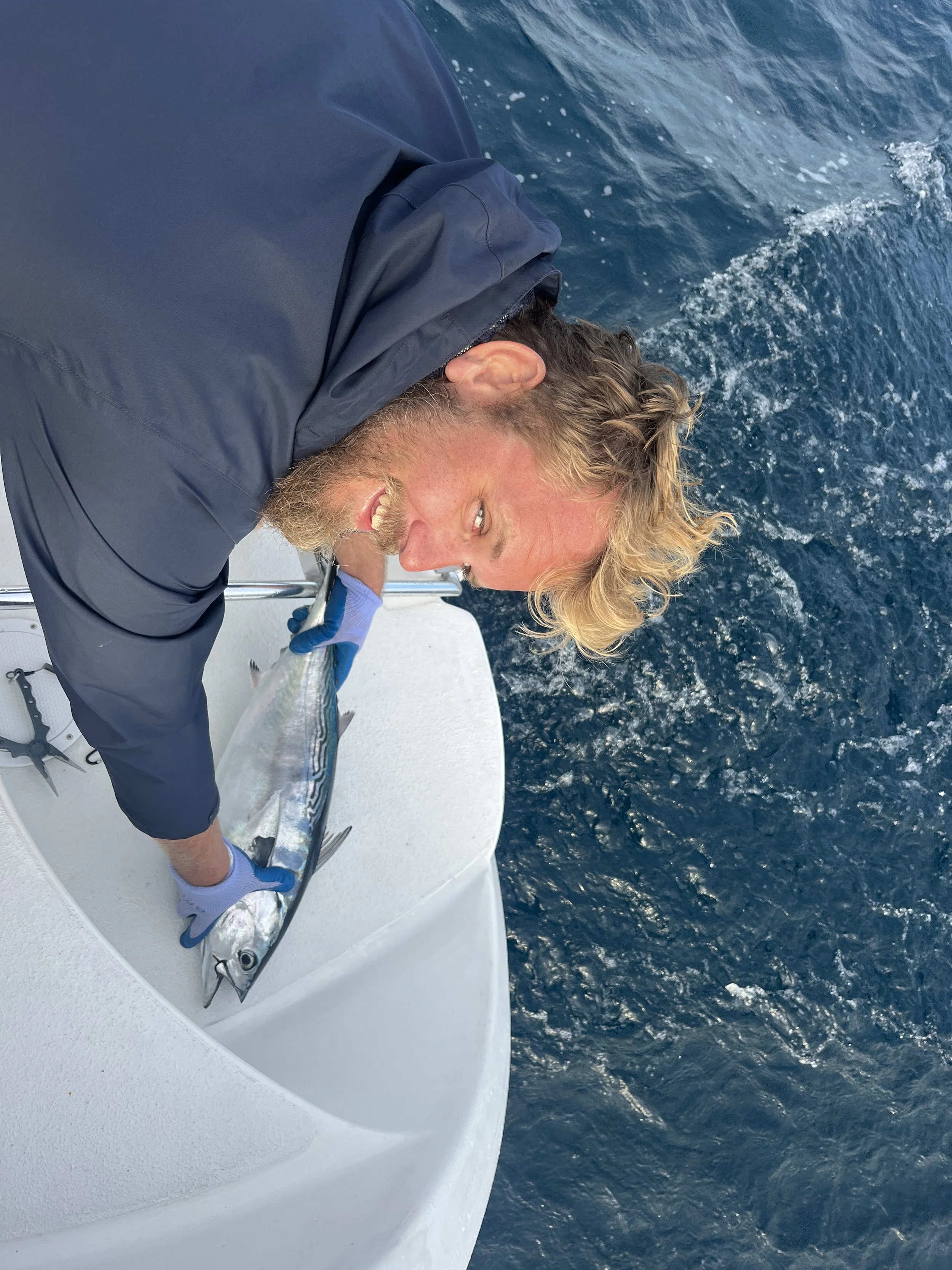 Man with blond hair and beard holding a fish on a boat in the water.
