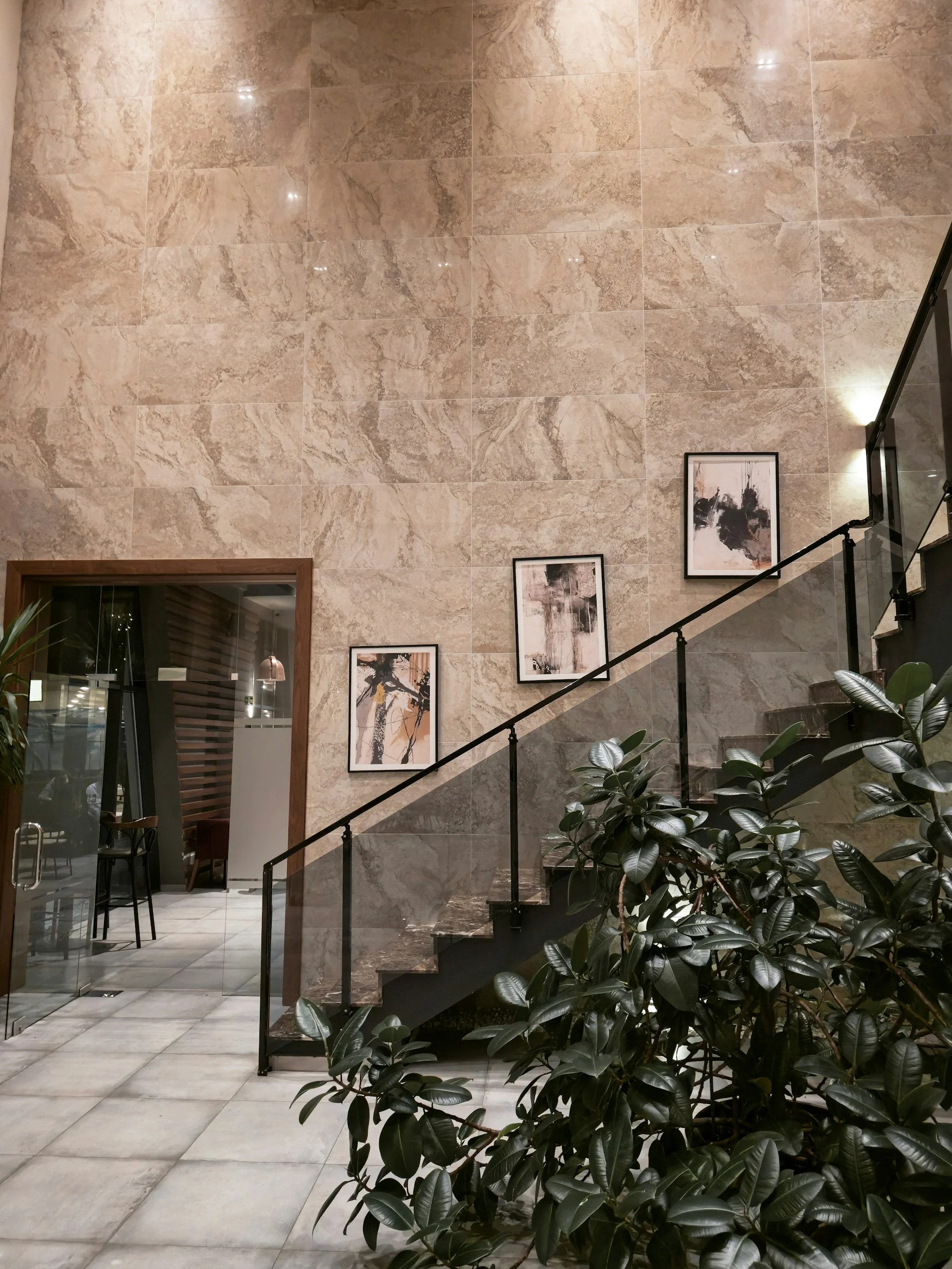 Interior view of a staircase area with beige marble walls, framed abstract artwork, and large green potted plant, with a glass door leading to another room.