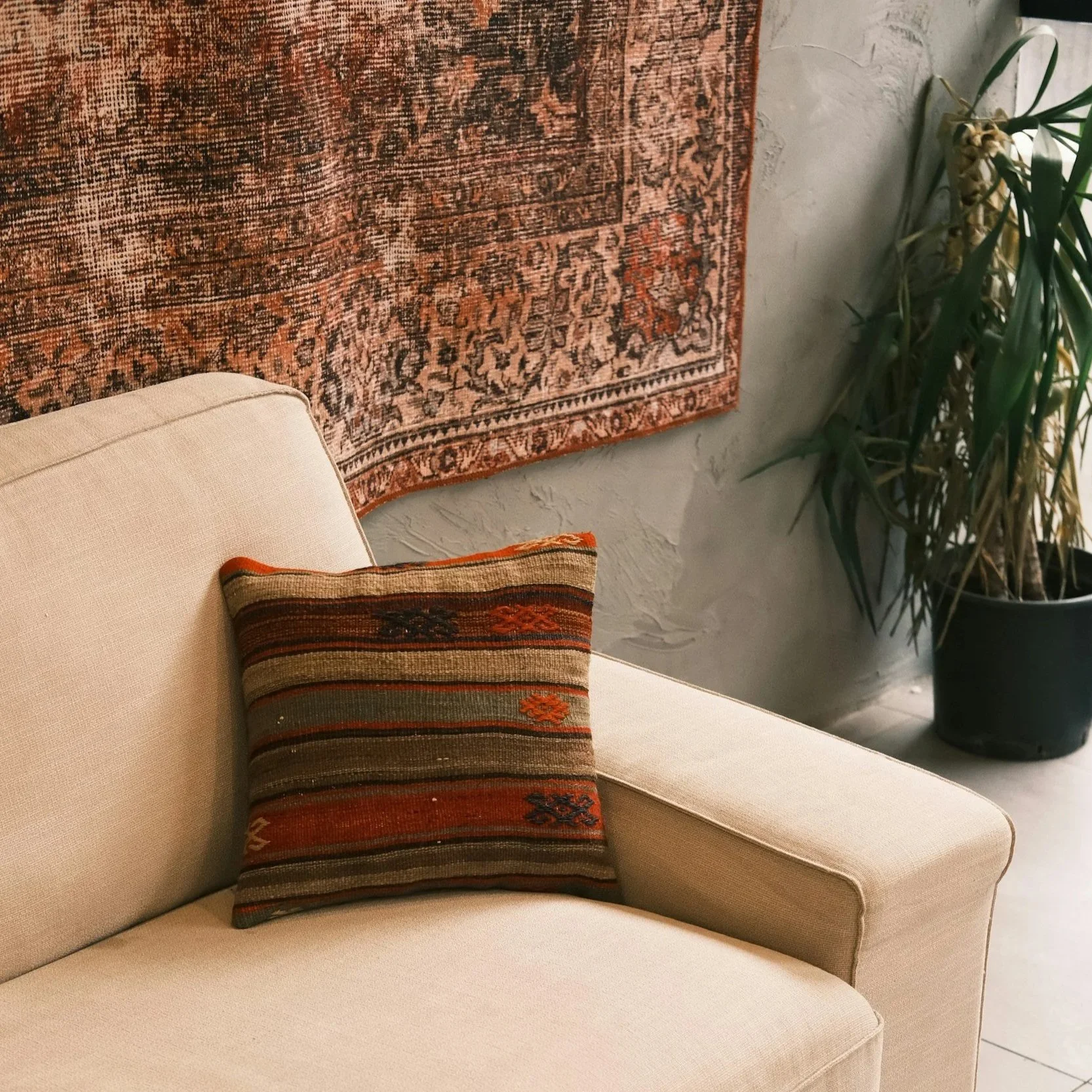A beige sofa with a decorative pillow that has a striped pattern with orange, green, and beige colors. Behind the sofa, there is a wall with a large, ornate, multicolored rug hanging on it and a tall potted plant with long, green leaves in a black pot.