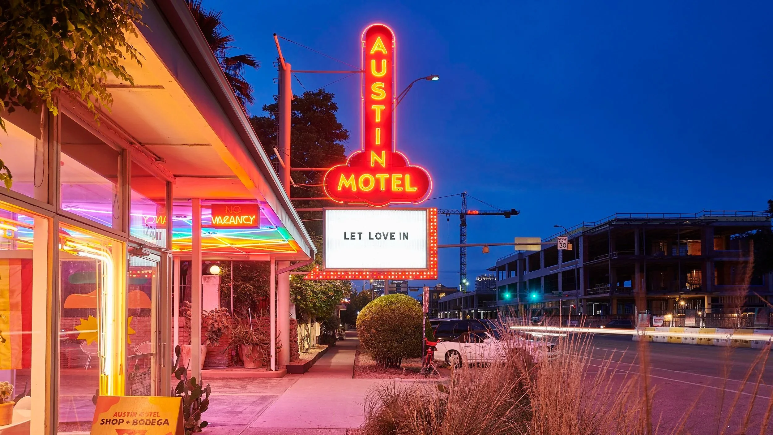 Neon sign for a motel that reads: 'AUSTIN MOTEL'. Below the sign, there is a marquee with the words 'LET LOVE IN'. The motel is in a city at dusk, with a construction crane, buildings, and street lights visible in the background. The building has colorful neon lighting, and there are parked cars and a small landscaped area in front.