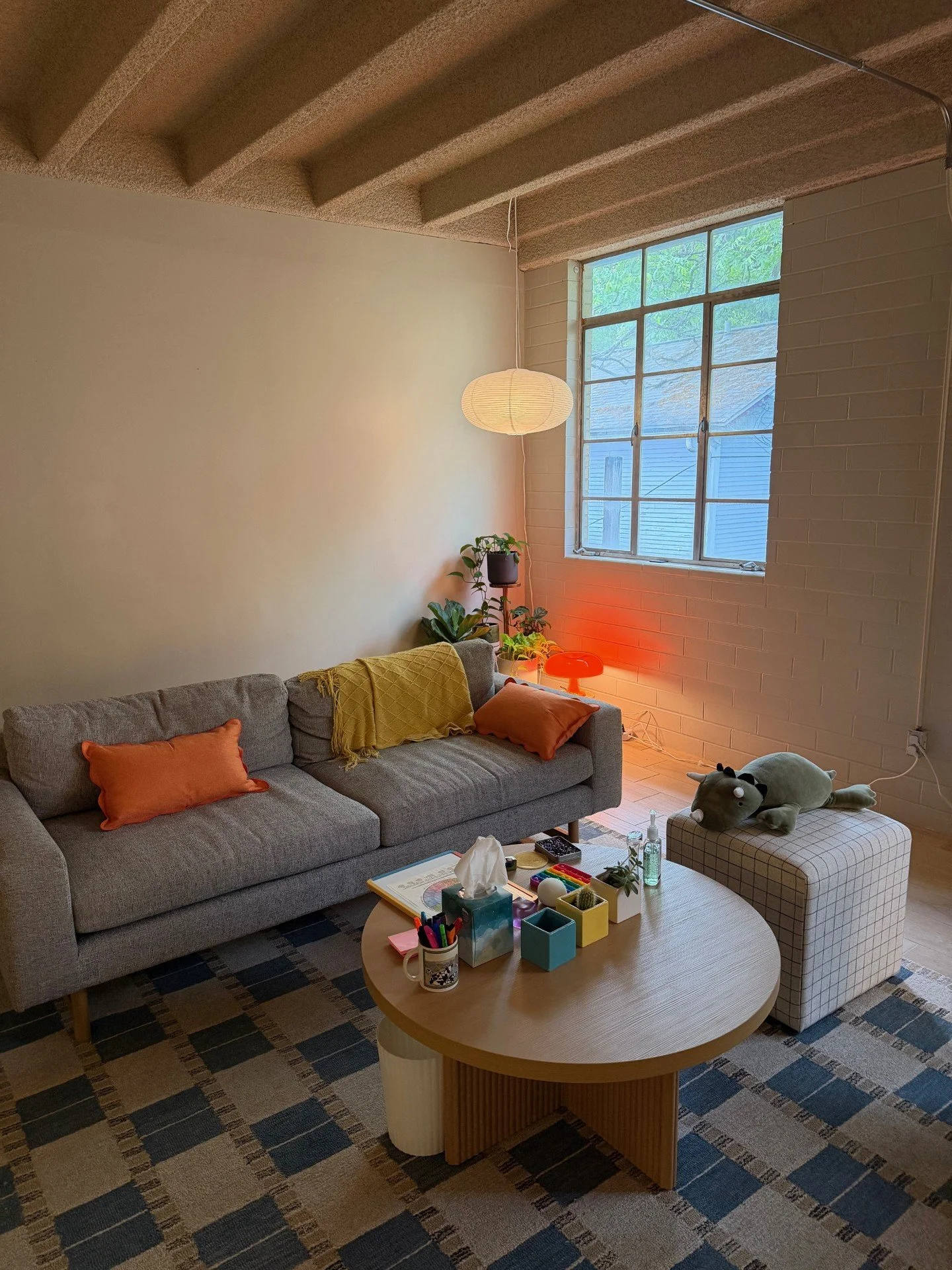 Living room with gray sofa, orange pillows, a yellow throw, a round wooden coffee table with various items, a checkered ottoman with a stuffed animal, a tall window, plants, a hanging paper lamp, a red side lamp, and a multicolored rug.