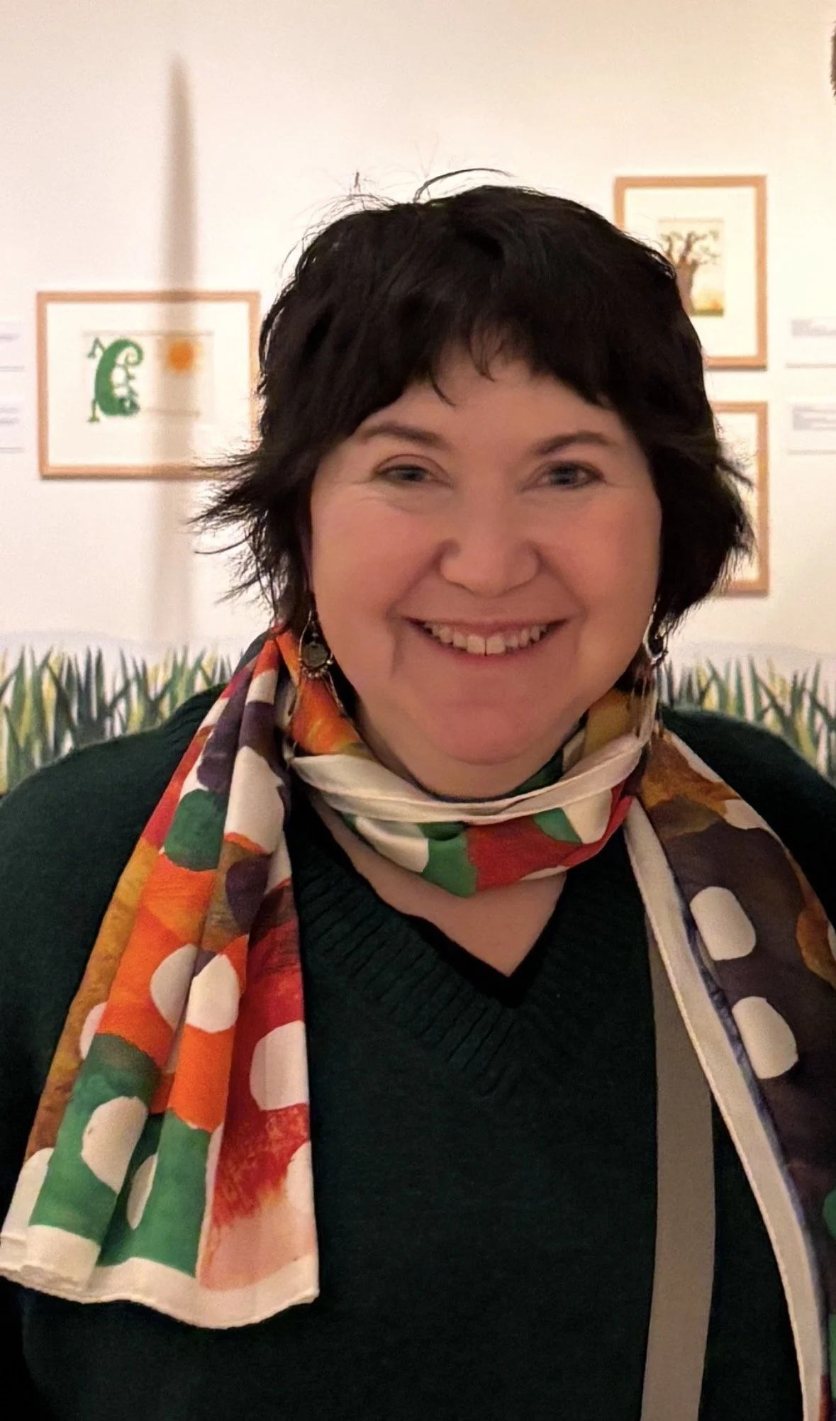 A woman with short dark hair smiling, wearing a colorful patterned scarf and a black top, in an art gallery with framed artwork on the wall behind her.