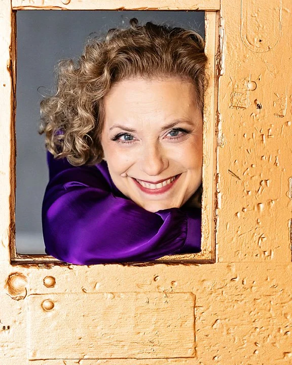 Woman with curly blonde hair smiling, looking through a small square window in a wooden wall, wearing a purple satin blouse.
