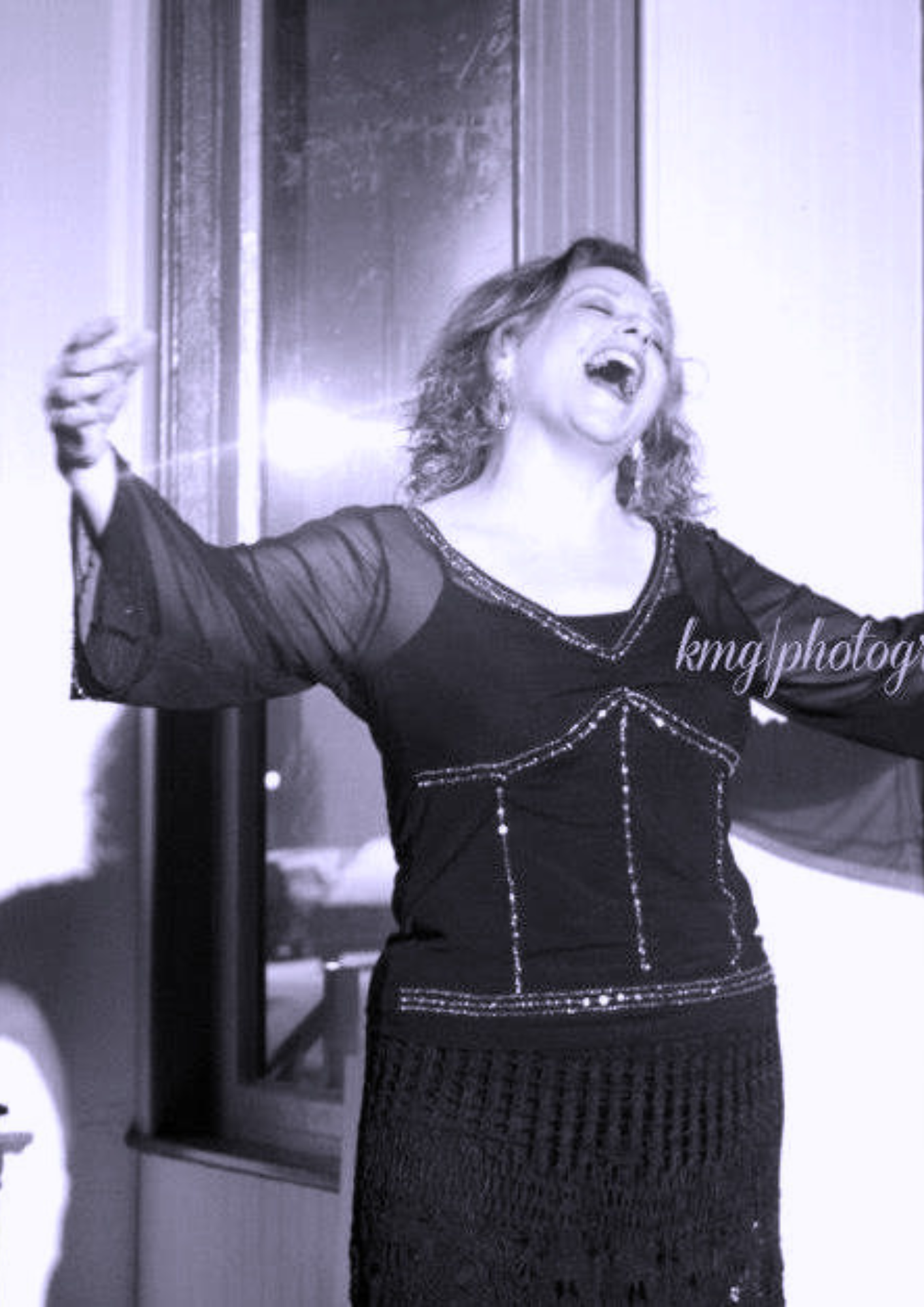 Woman smiling and laughing with arms outstretched, wearing a black dress with decorative stitching, standing indoors near a mirror.